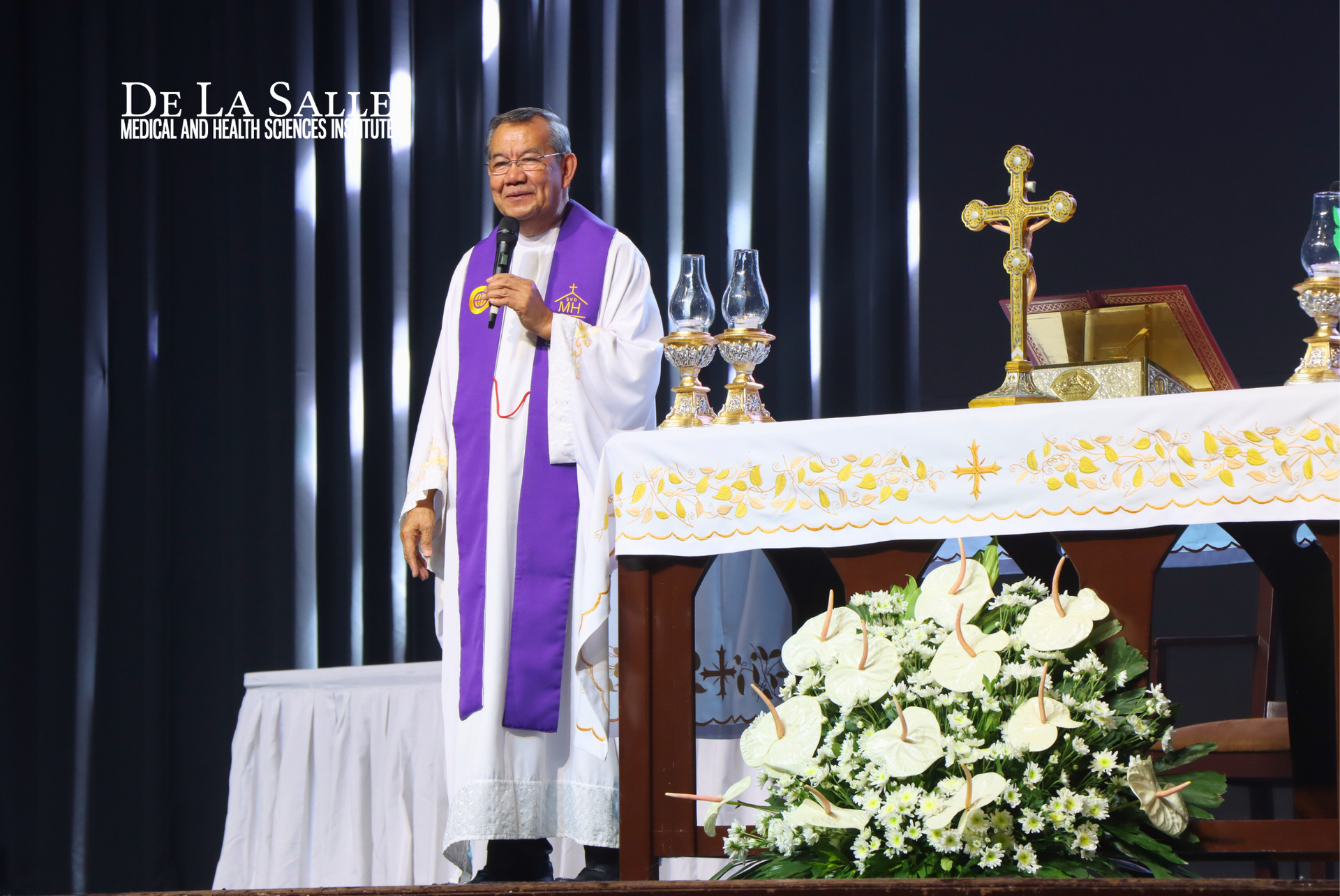 Priest in white and purple vestments stands beside an altar with flowers and a gold cross during a church service