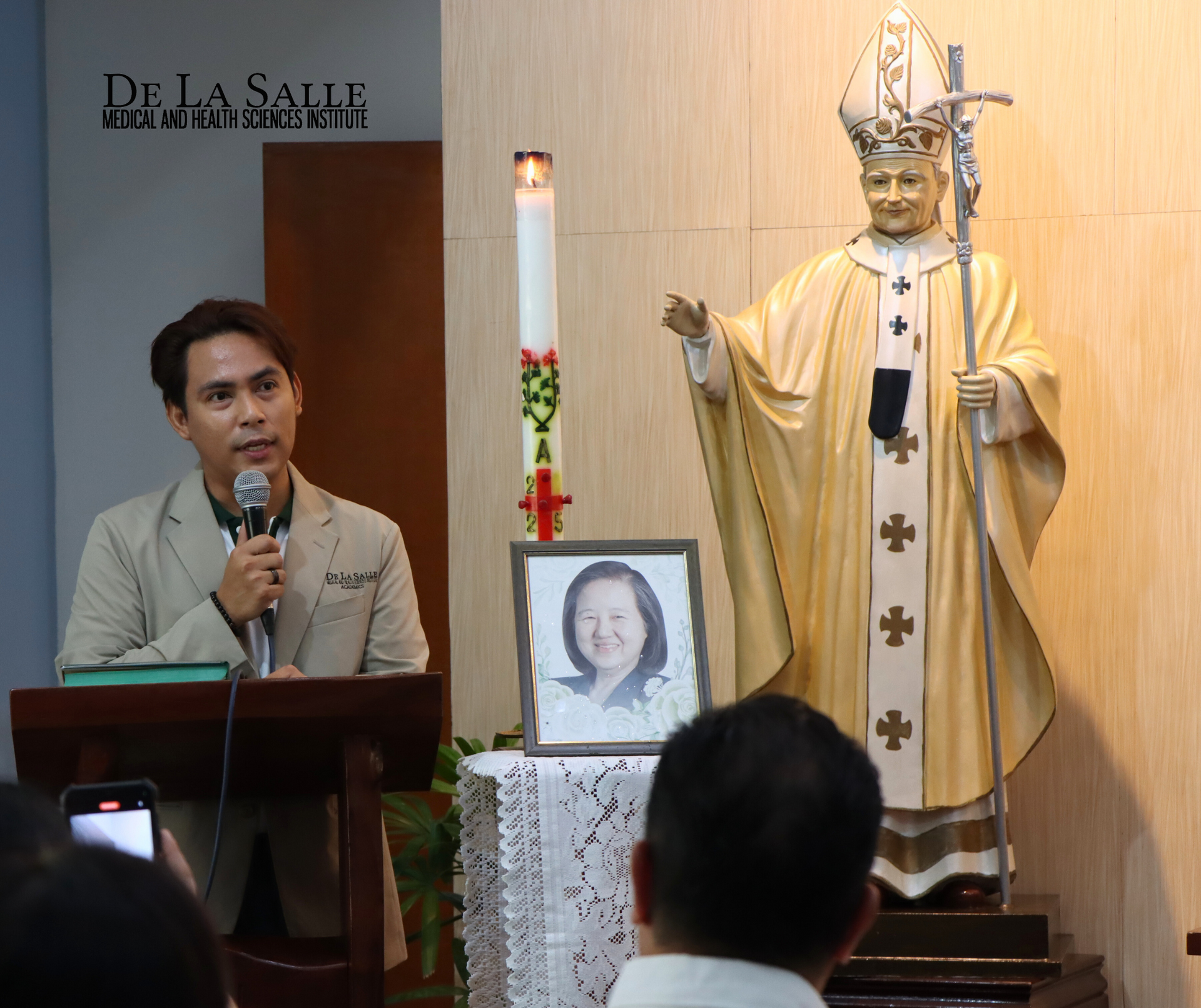 Man speaking at a podium beside a religious statue and framed portrait in a chapel setting