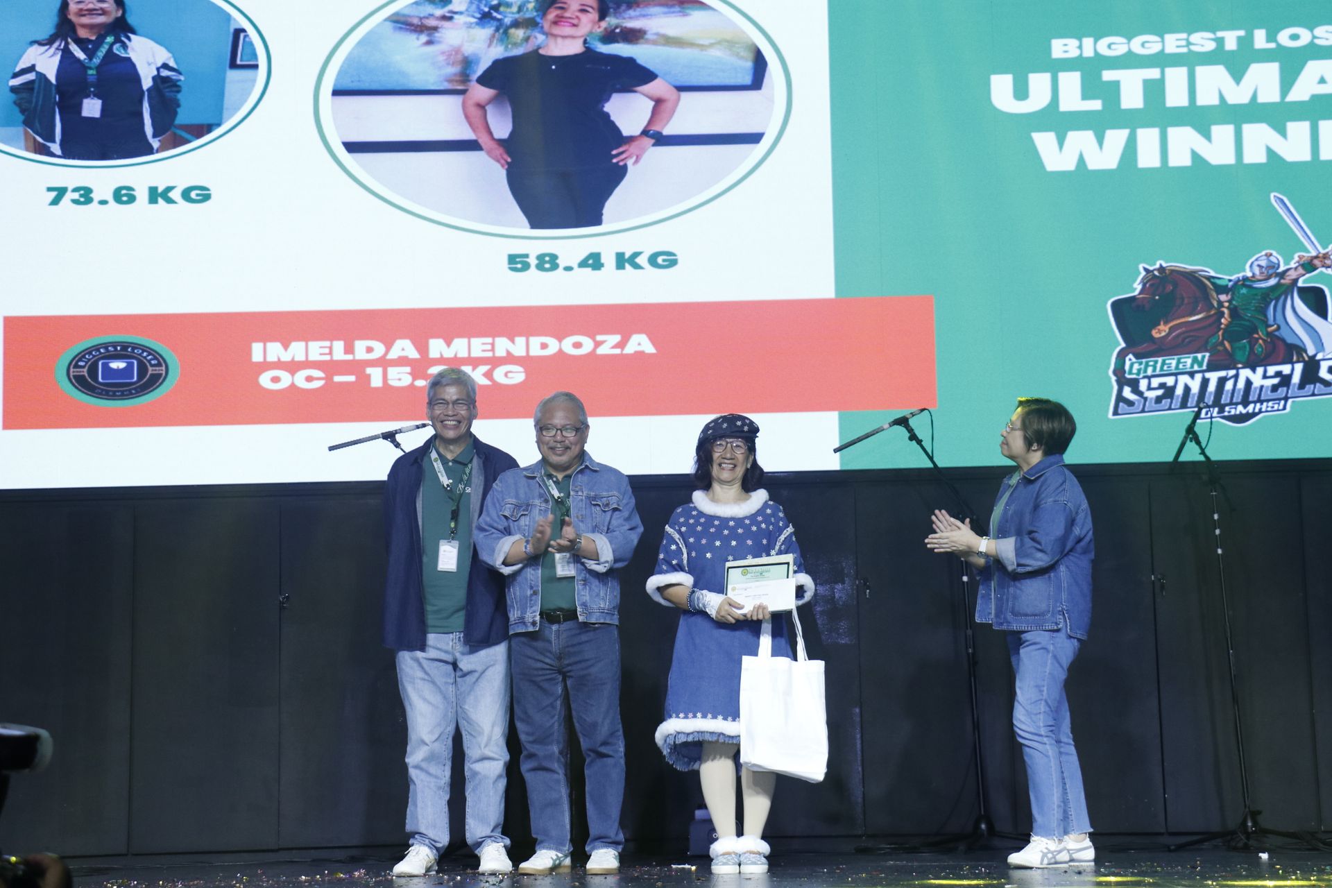 Four people on a stage during an awards ceremony, with a large event screen behind them.