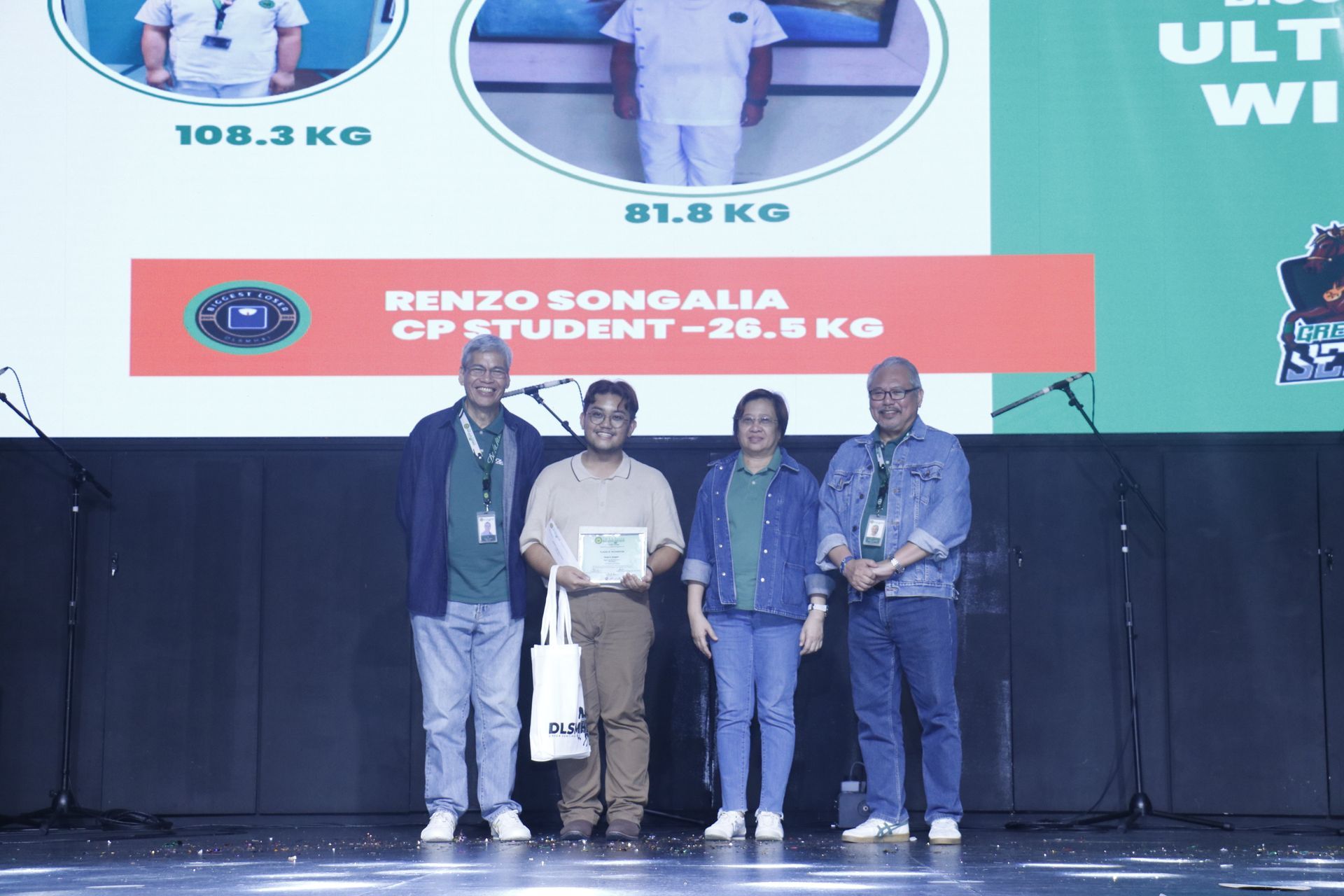 Four people on stage holding a certificate and gift bag beneath a sports event banner