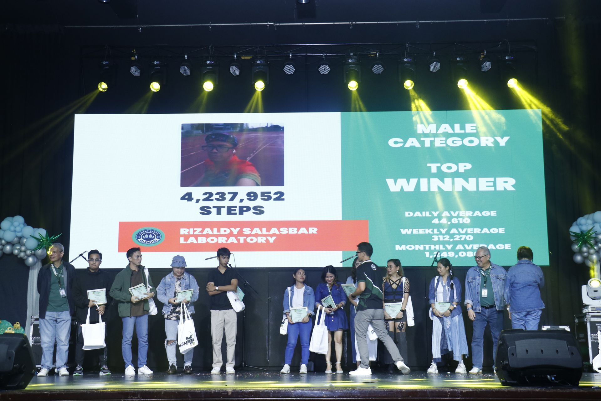 Award stage with a large group of people, trophies, and a screen announcing top winners under bright lights