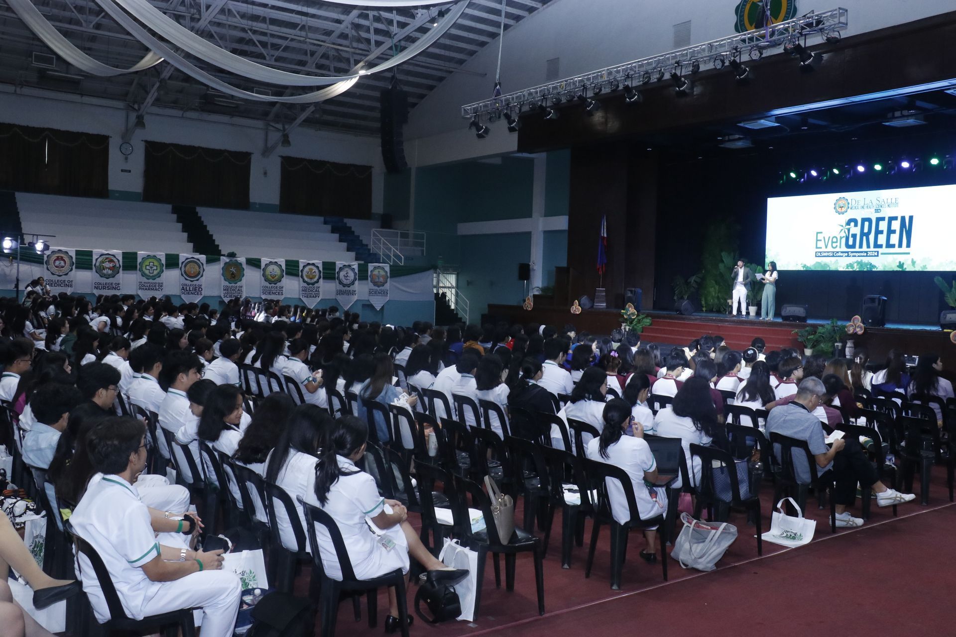 Students in white shirts seated in a large auditorium facing a stage with a green presentation screen