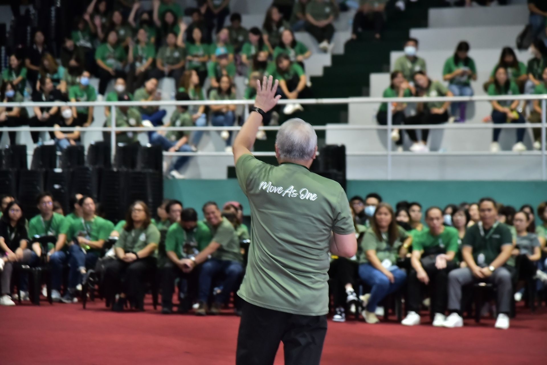 Coach in green shirt raises hand on a badminton court before a seated crowd in green shirts.