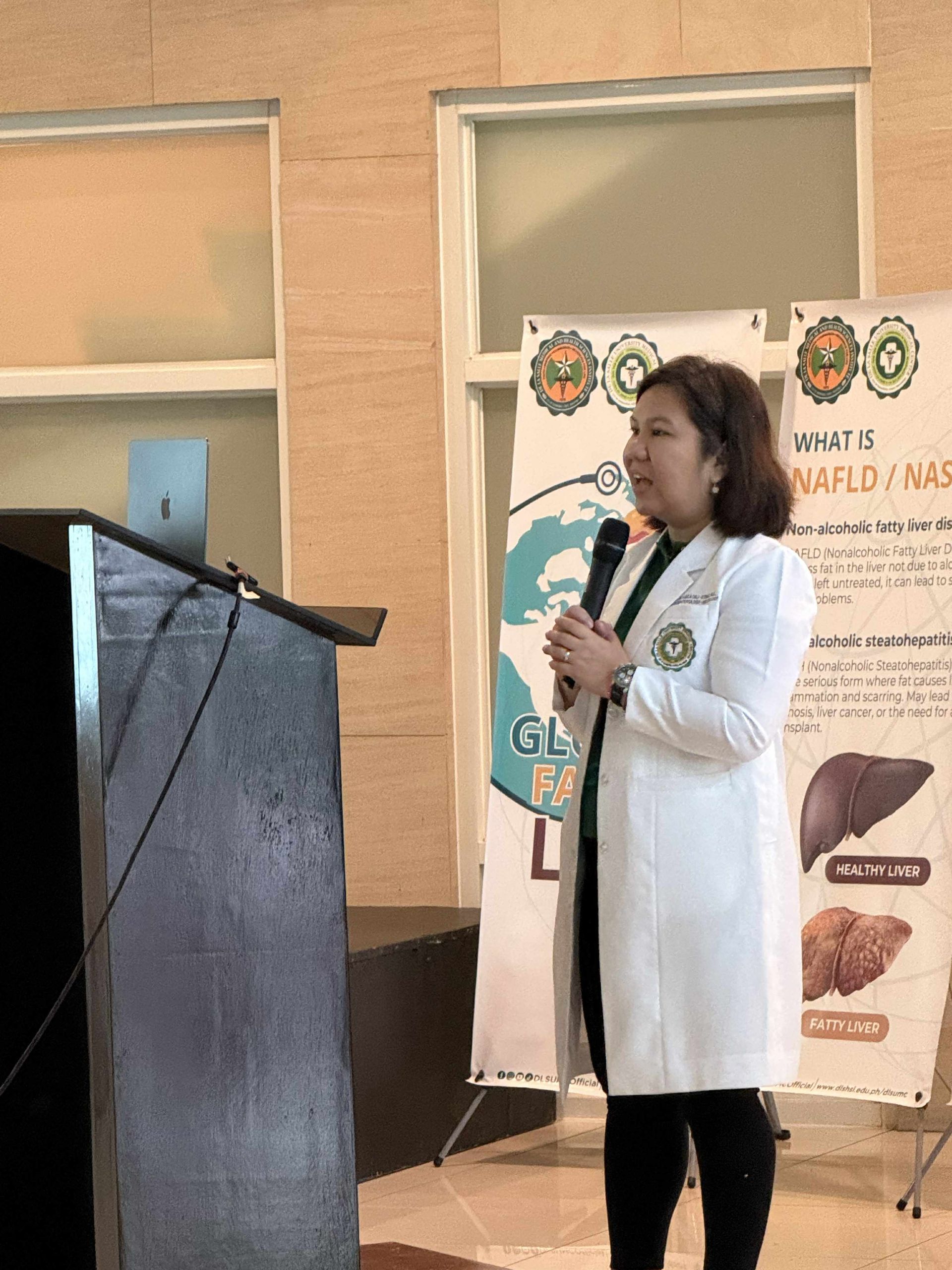 Woman speaking at a podium during a presentation beside health posters and a banner.