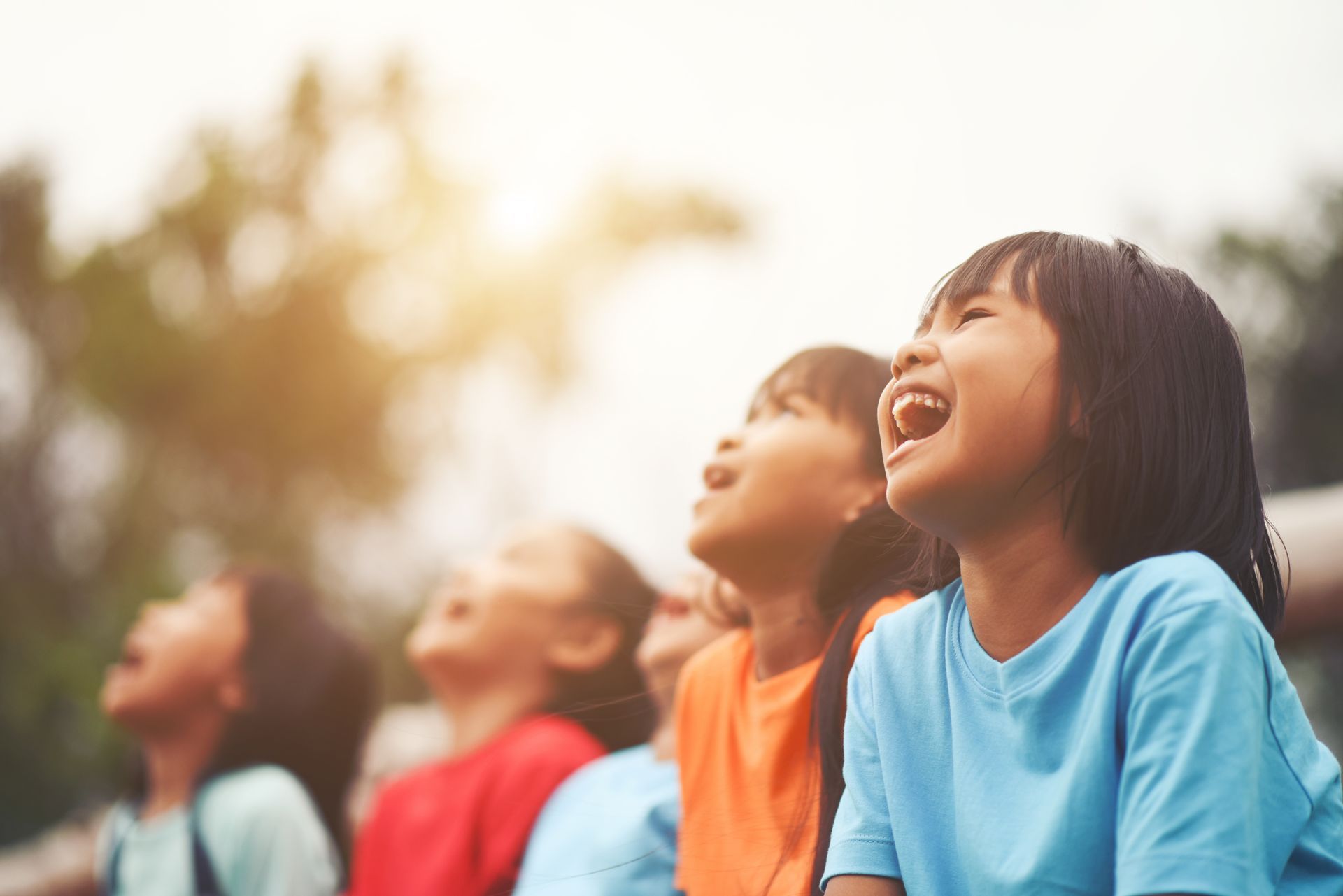 Children laughing outdoors in colorful shirts, looking up in bright sunlight