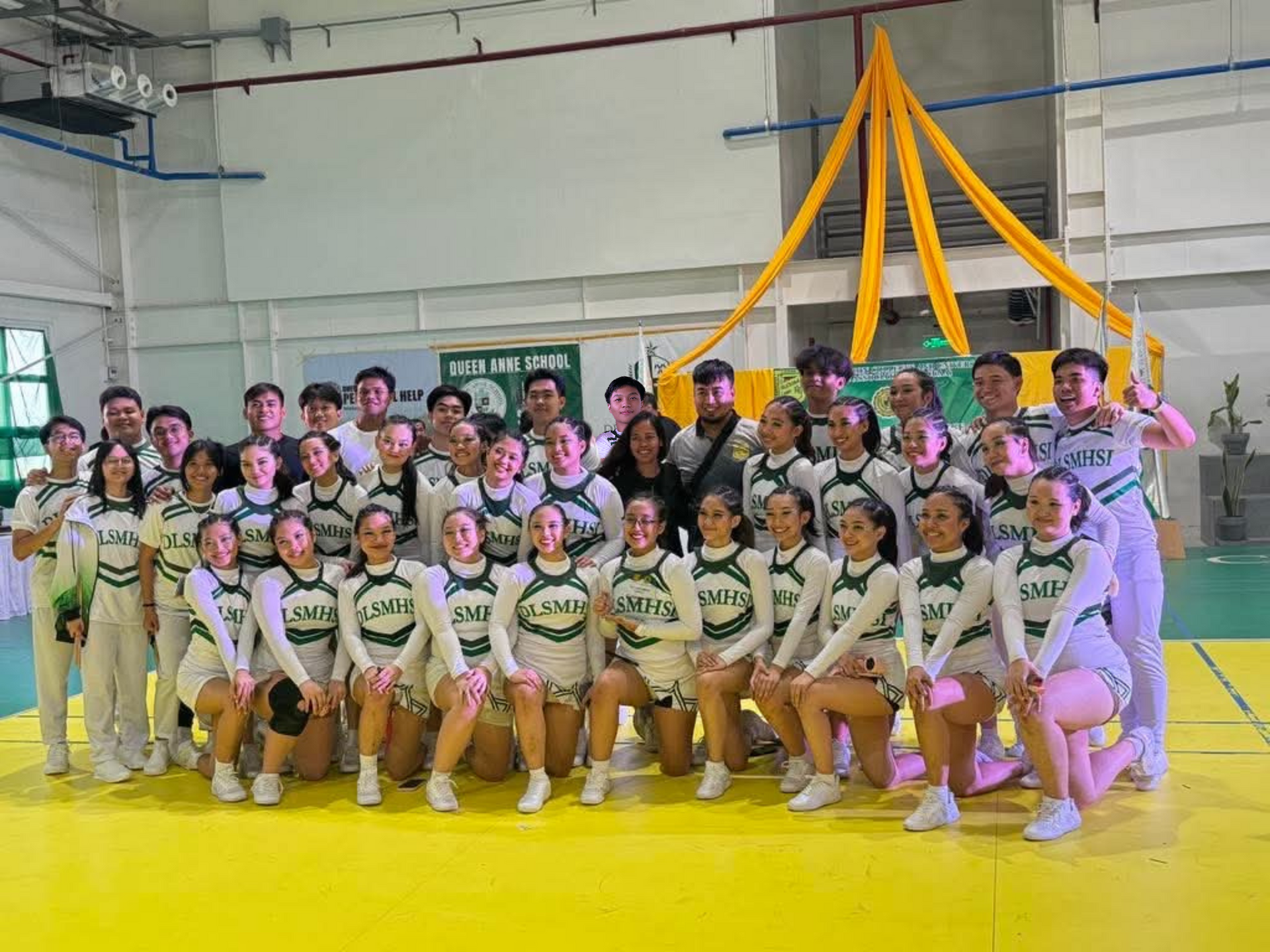 Group of cheerleaders posing in a gym, wearing white and pink uniforms with a yellow ribbon backdrop