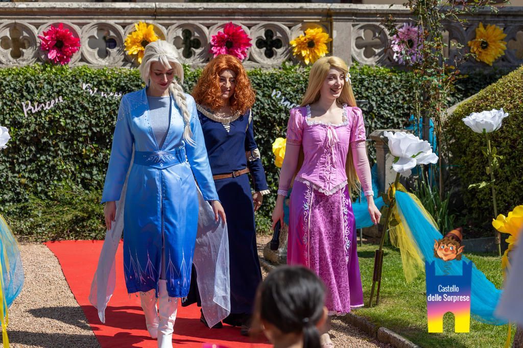 Tre artiste vestite da Elsa, Merida e Rapunzel sfilano su un tappeto rosso all'aperto, tra colorate decorazioni floreali.