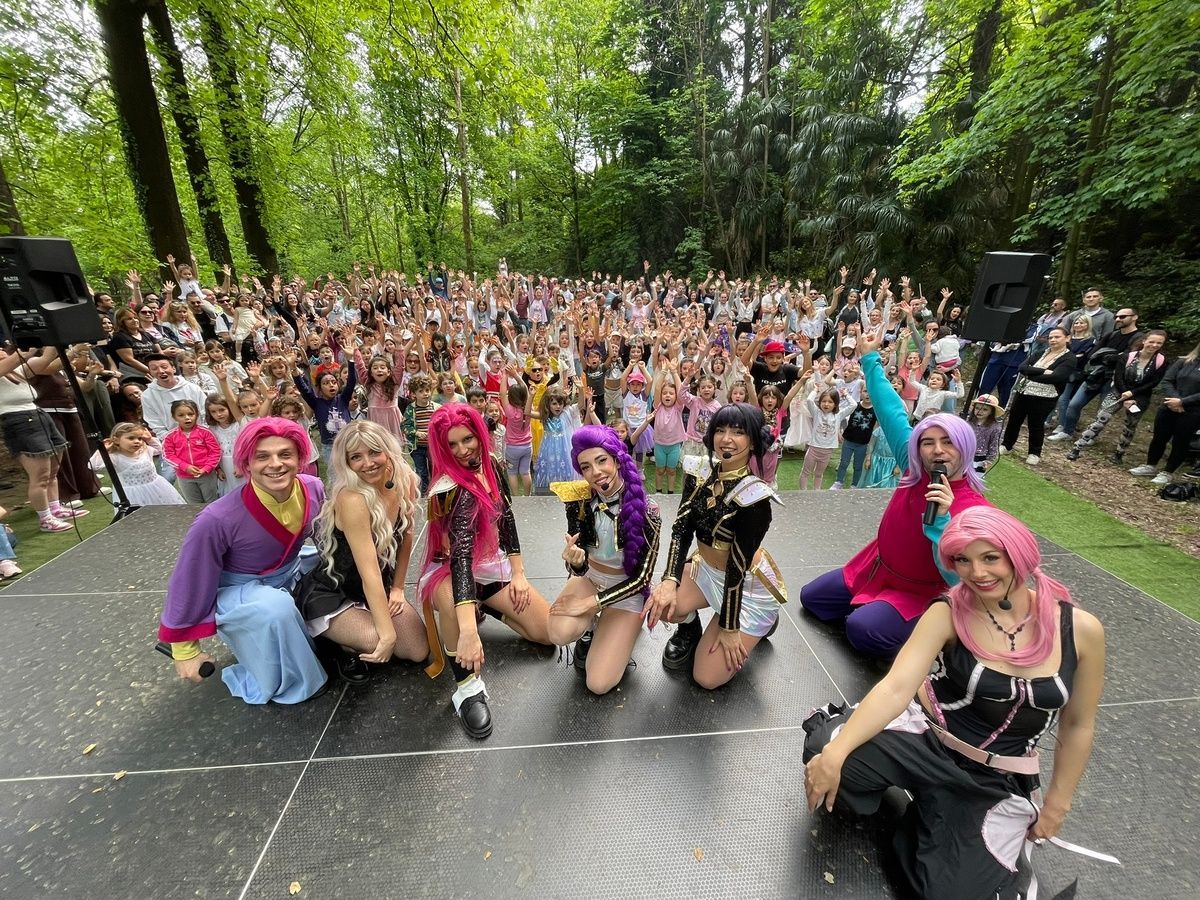 Un gruppo di ballerini vestiti con abiti colorati posa all'aperto in un giardino, con un cartello rosa con la scritta