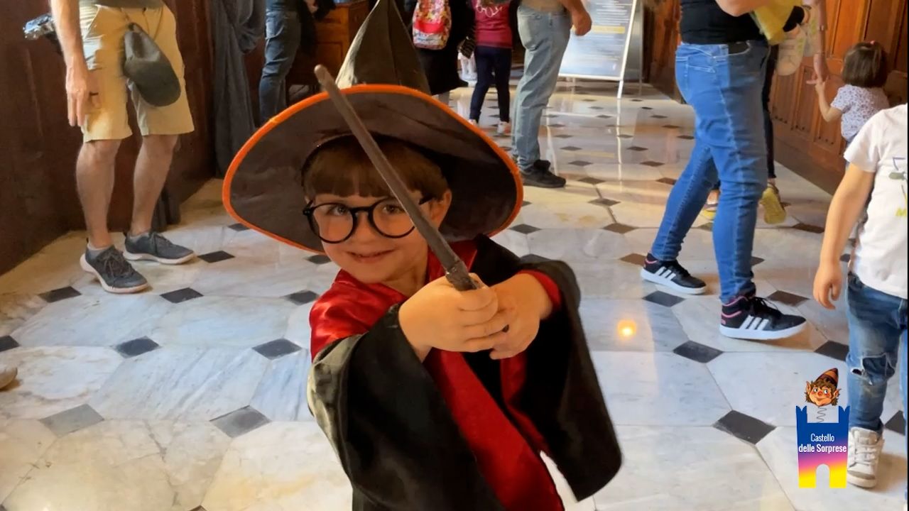 Un bambino piccolo, vestito da mago con tunica, cappello e occhiali, tiene giocosamente in mano una bacchetta magica in uno spazio interno piastrellato.