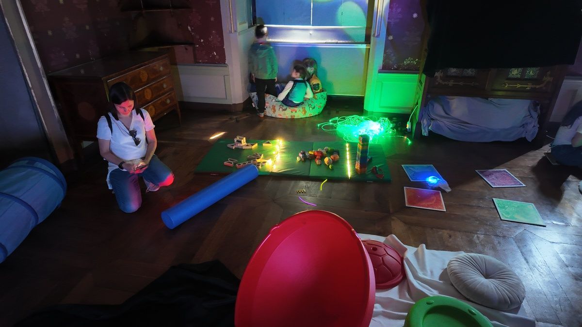 Un gruppo di bambini e adulti siede sul pavimento in una stanza sensoriale scarsamente illuminata da luci a fibra ottica colorate e brillanti.