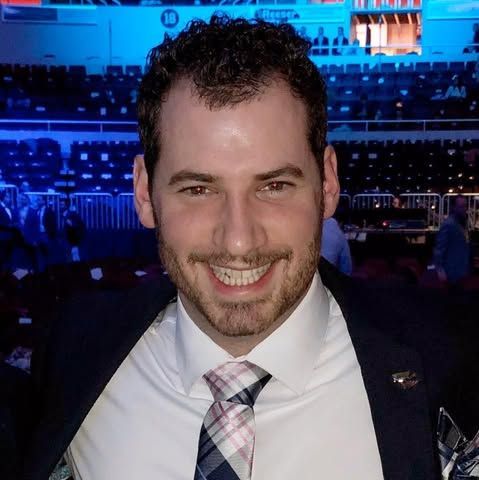Man in white shirt and patterned tie smiles in arena setting.