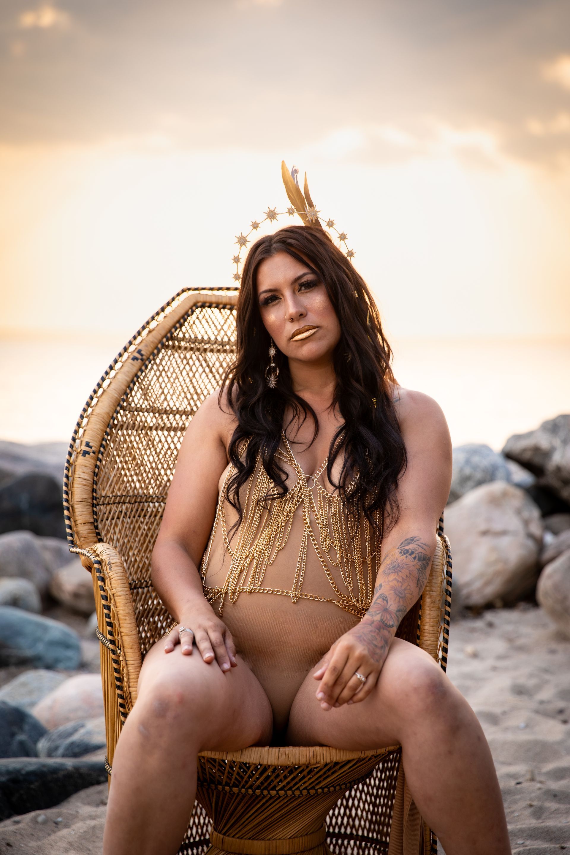 Woman in a gold dress seated on a wicker chair at a rocky beach during sunset