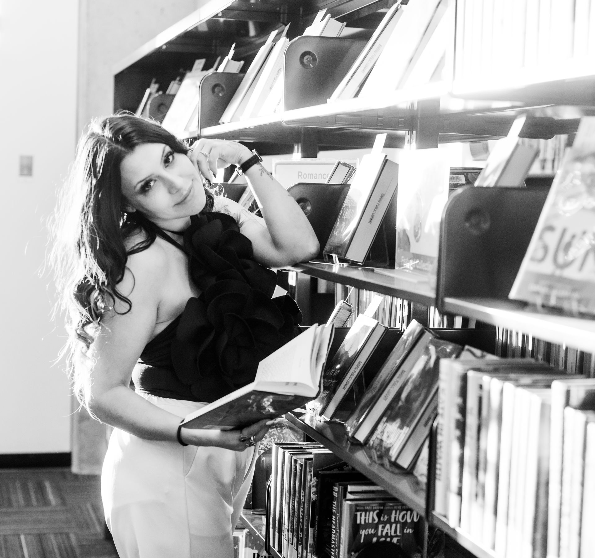 Person posing in a bookstore aisle, leaning toward shelves and holding a book, in black and white.