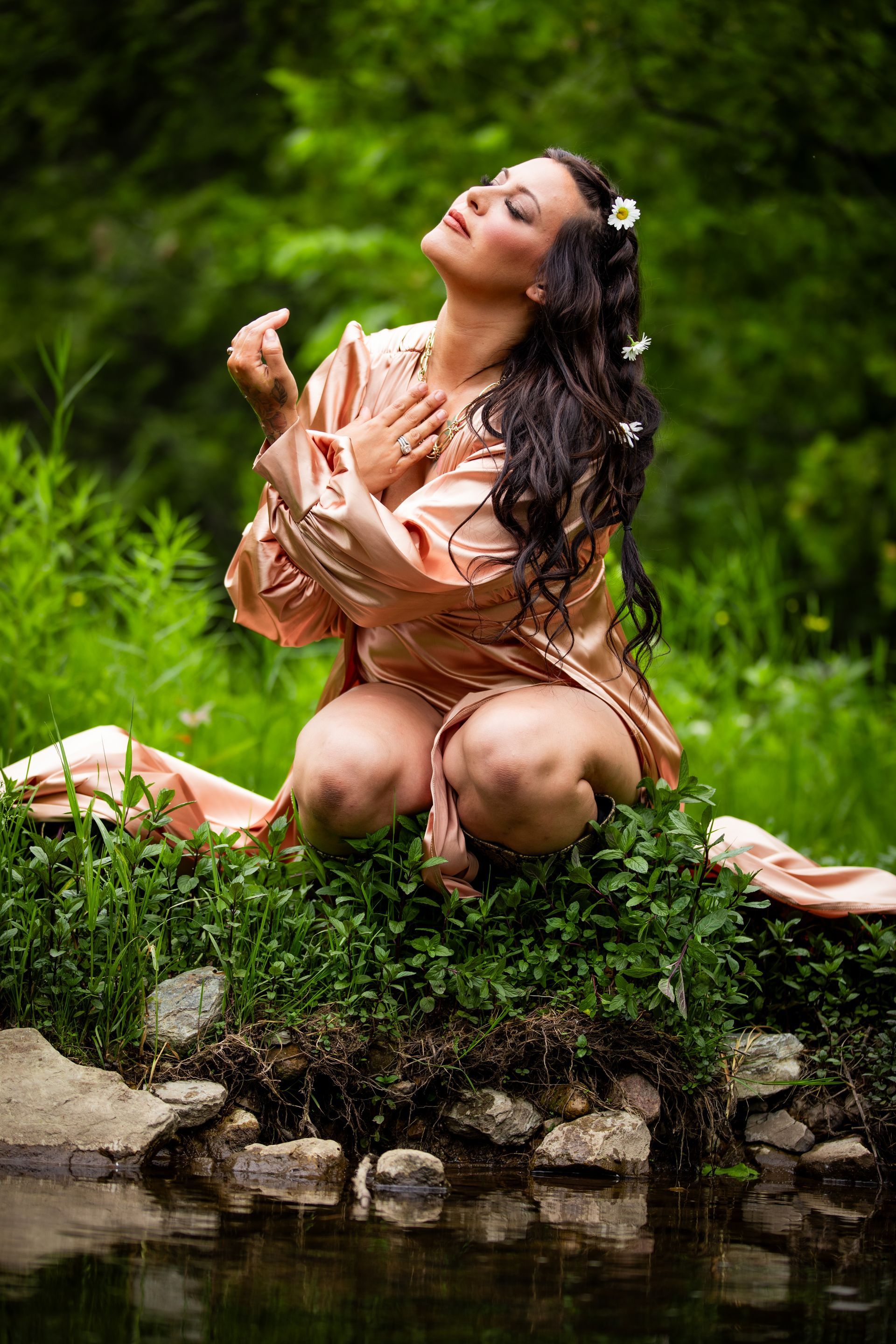 Woman in a peach dress kneeling by a pond, eyes closed, surrounded by lush green foliage.