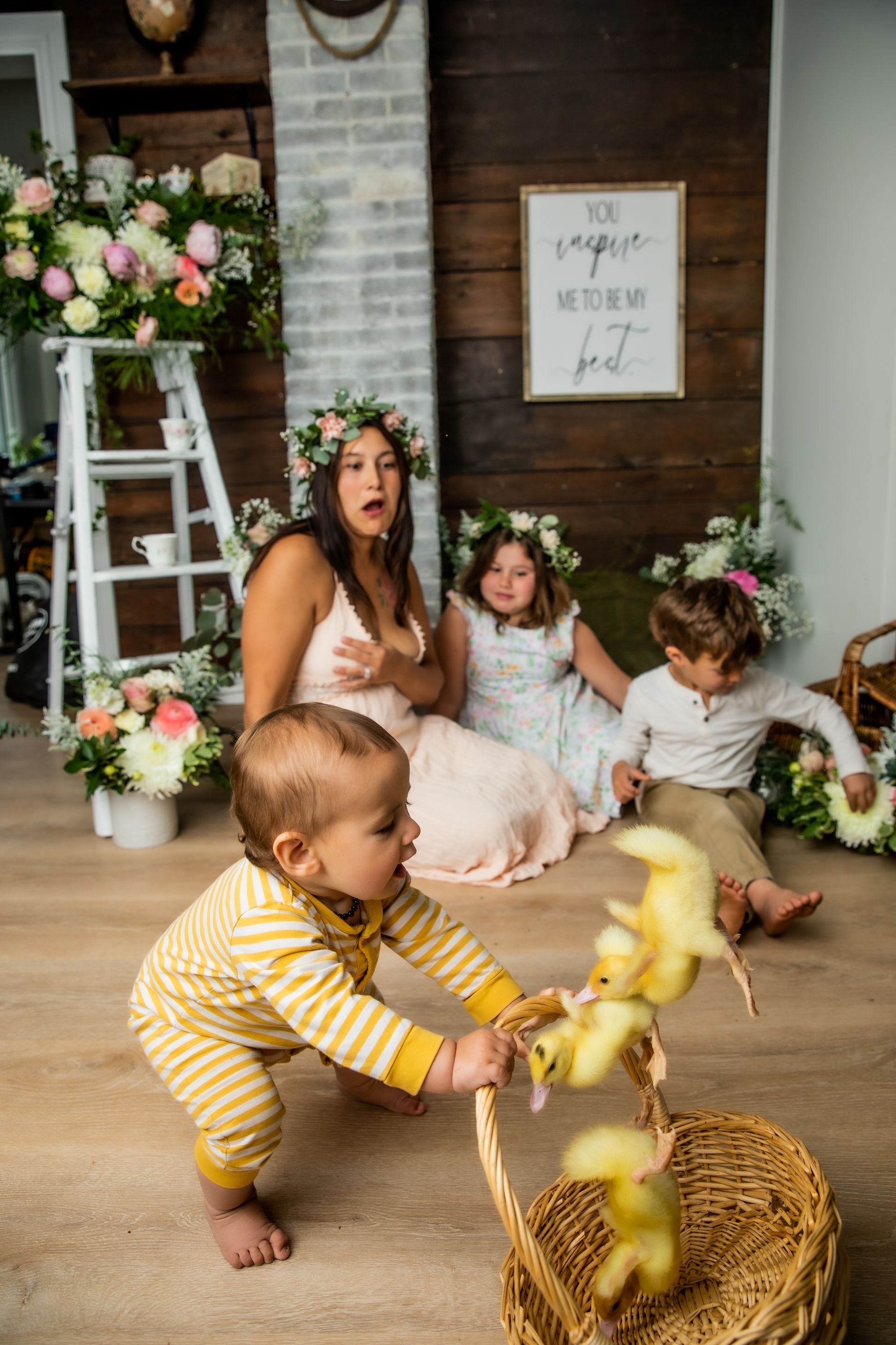 Woman and three children in a rustic floral nursery, with a toddler reaching for hanging yellow decorations.
