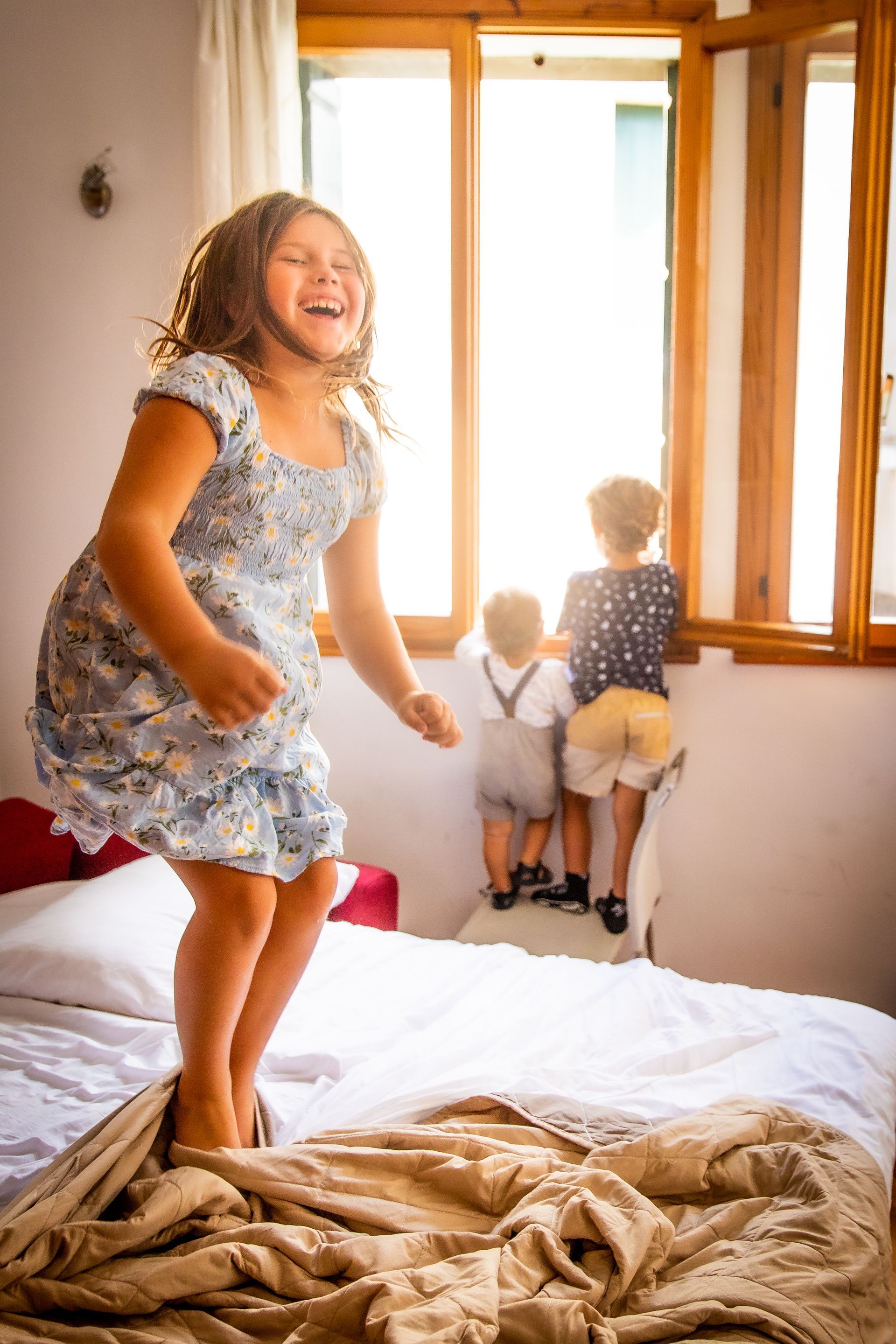 Children jumping on a bed in a sunlit bedroom near a window