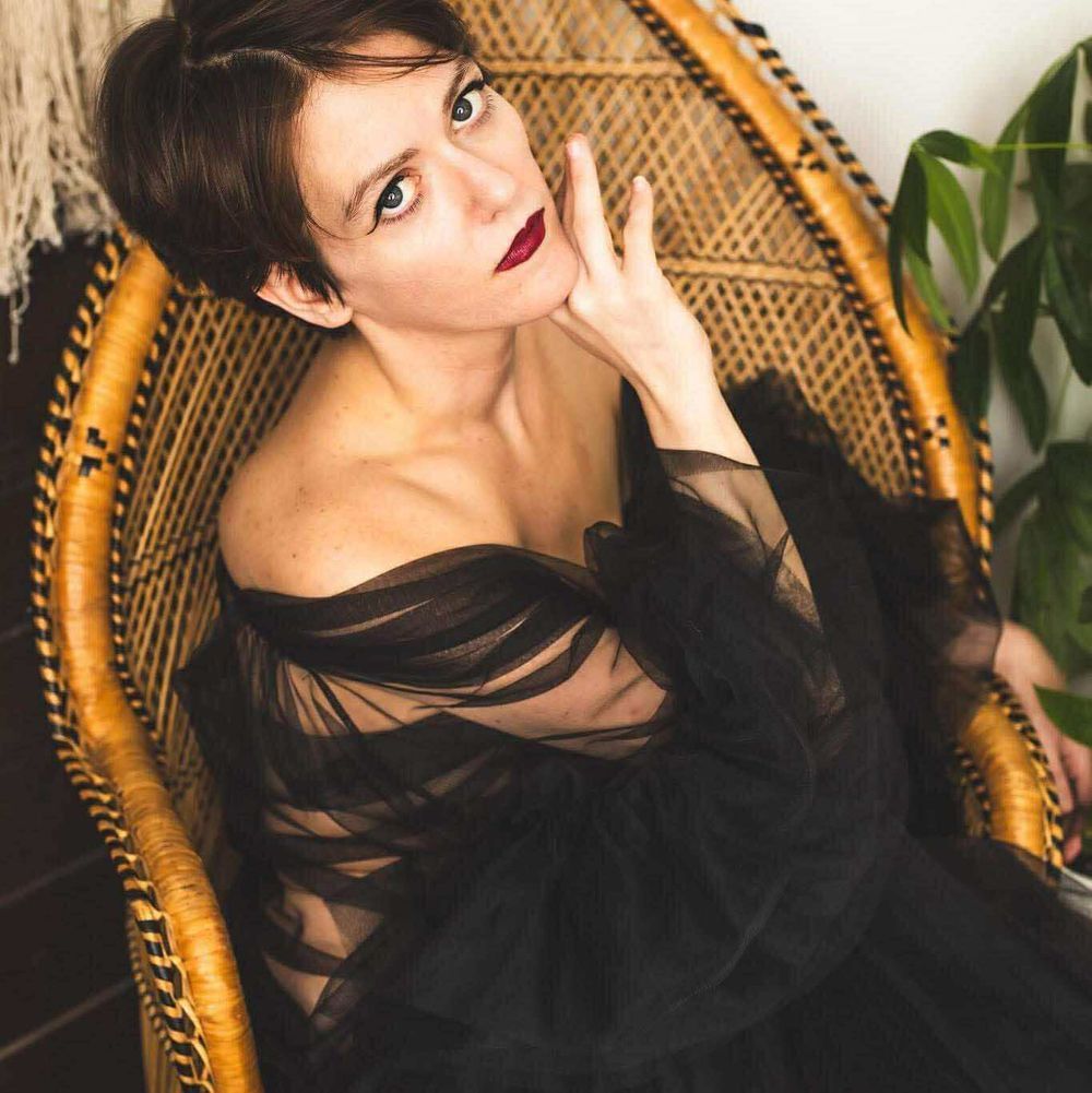 Person in a black sheer dress sitting in a wicker chair, looking at the camera with a hand near their neck.