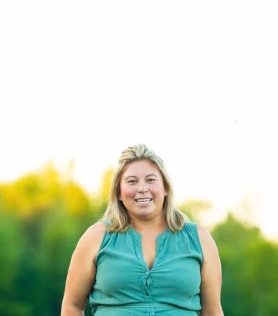 Smiling woman in a teal sleeveless top standing outdoors with blurred green and yellow foliage behind her