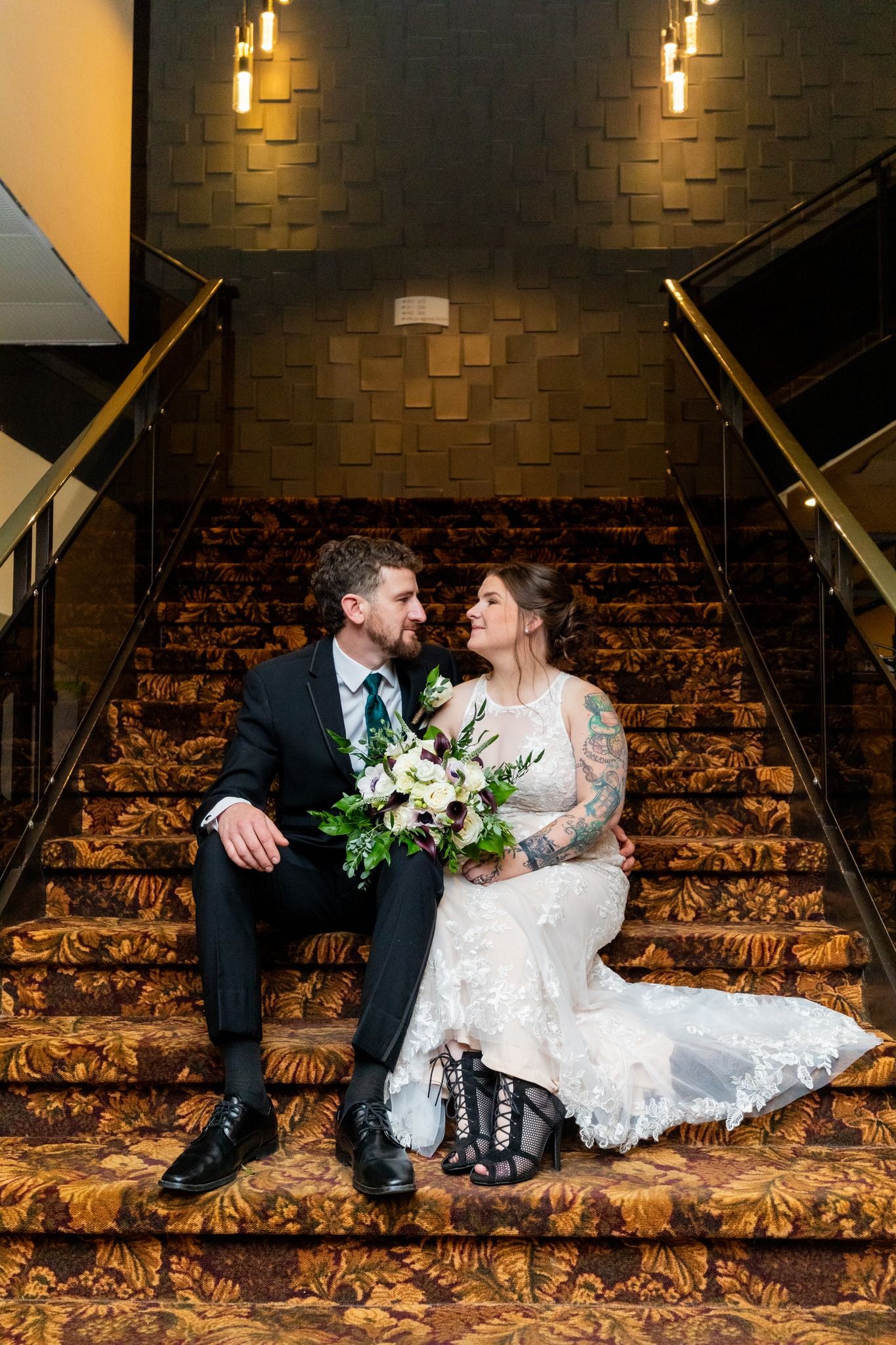 Bride and groom sitting on a staircase, smiling and holding a bouquet in a warmly lit venue
