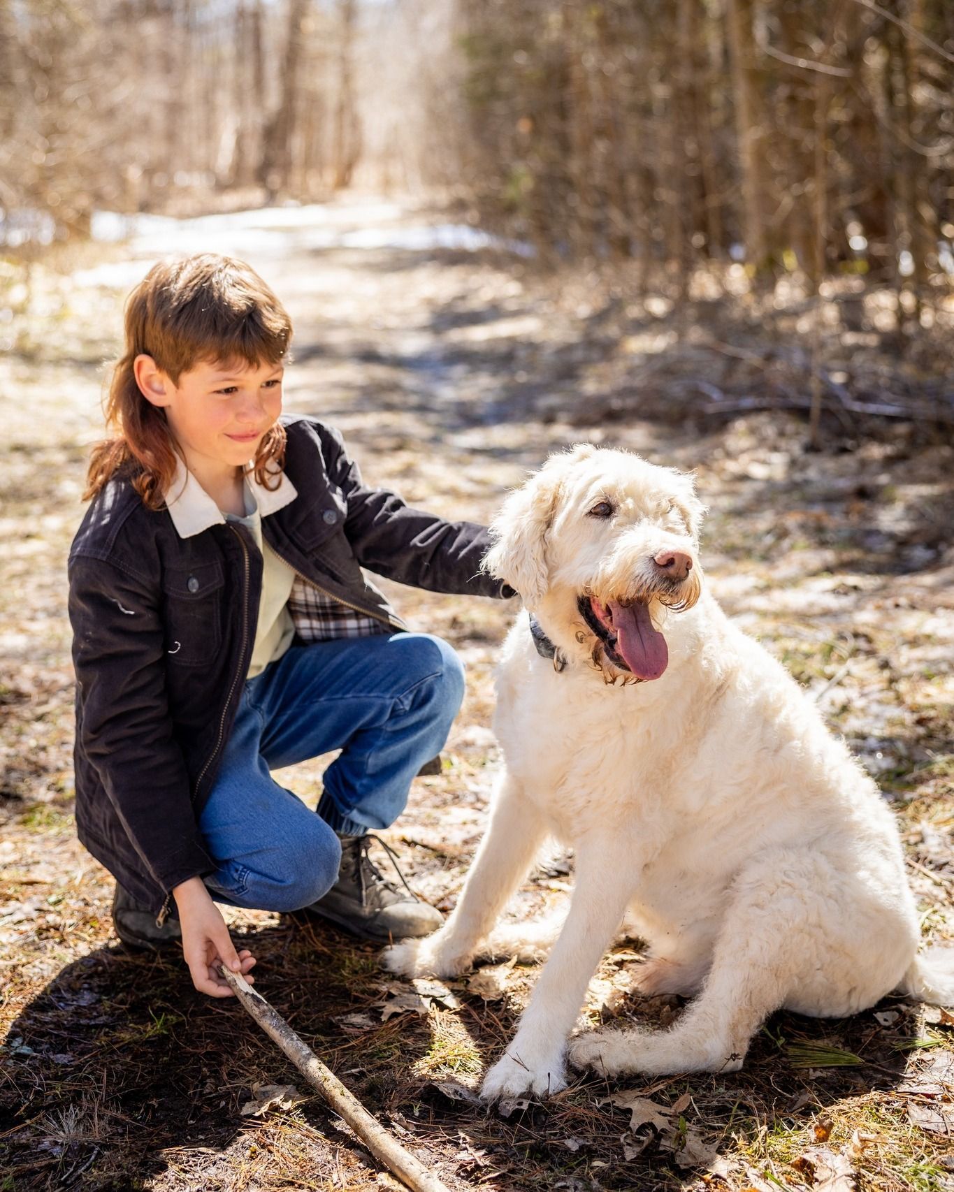 Boy kneeling beside a fluffy white dog on a wooded path in sunlight