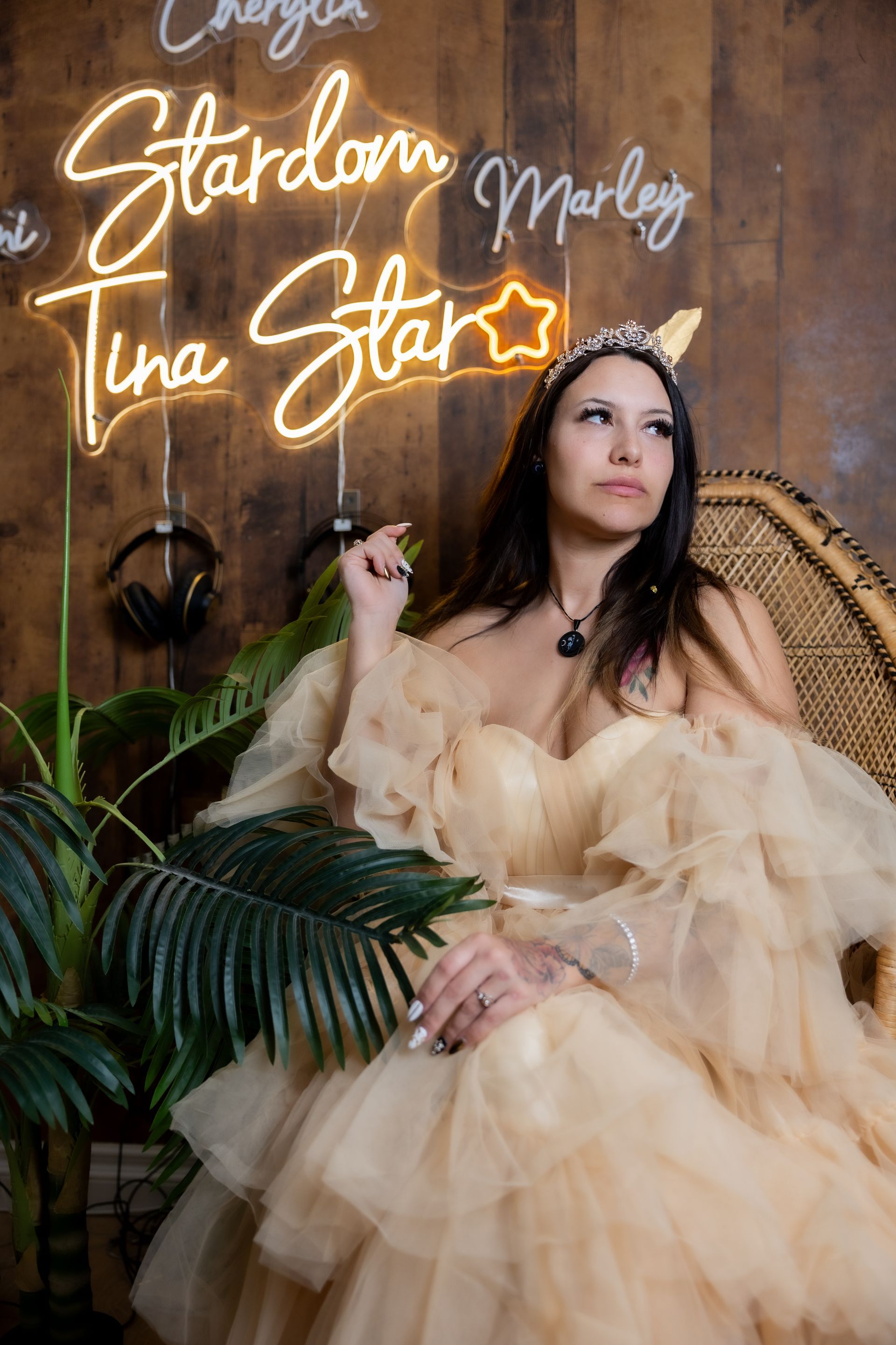 Woman in a cream tulle dress seated against a wood wall with neon names and stars, framed by greenery