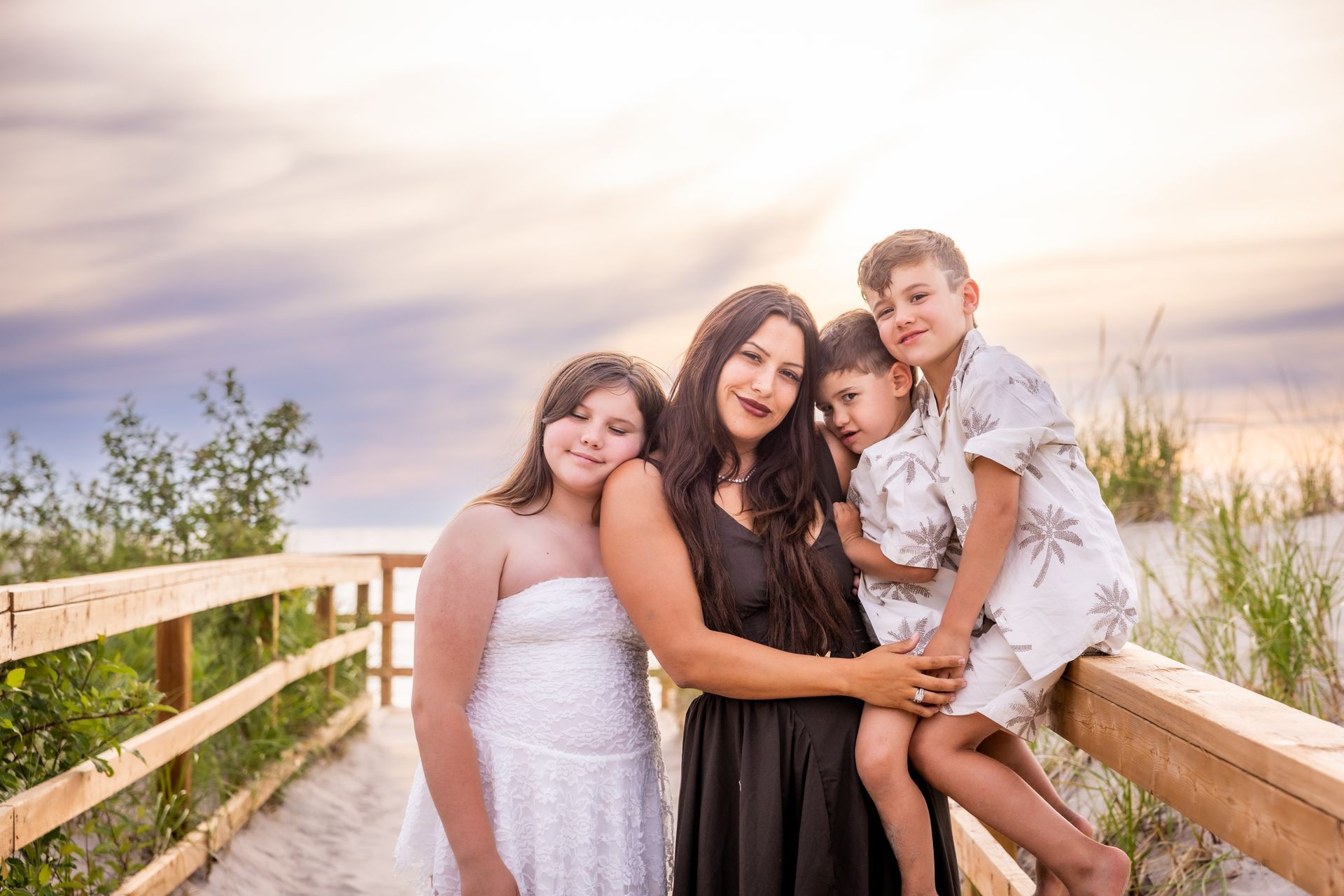 Four people leaning on a wooden boardwalk railing at sunset, with beach grass and a soft sky behind them