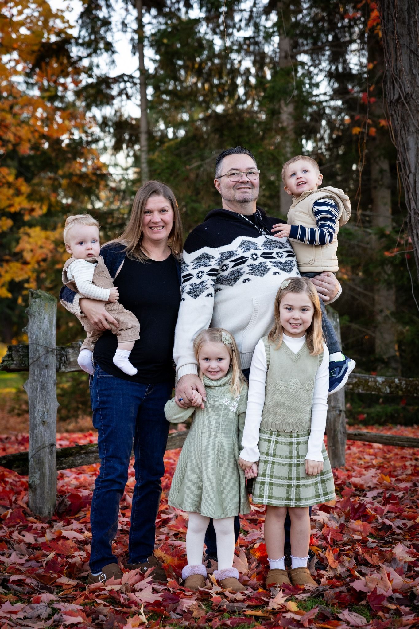 Family of five posing outdoors in autumn woods, with red leaves and trees in the background.