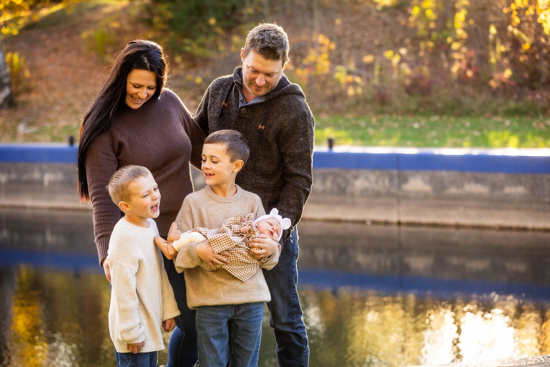 Family standing by a canal, smiling and admiring a newborn baby in a blanket.