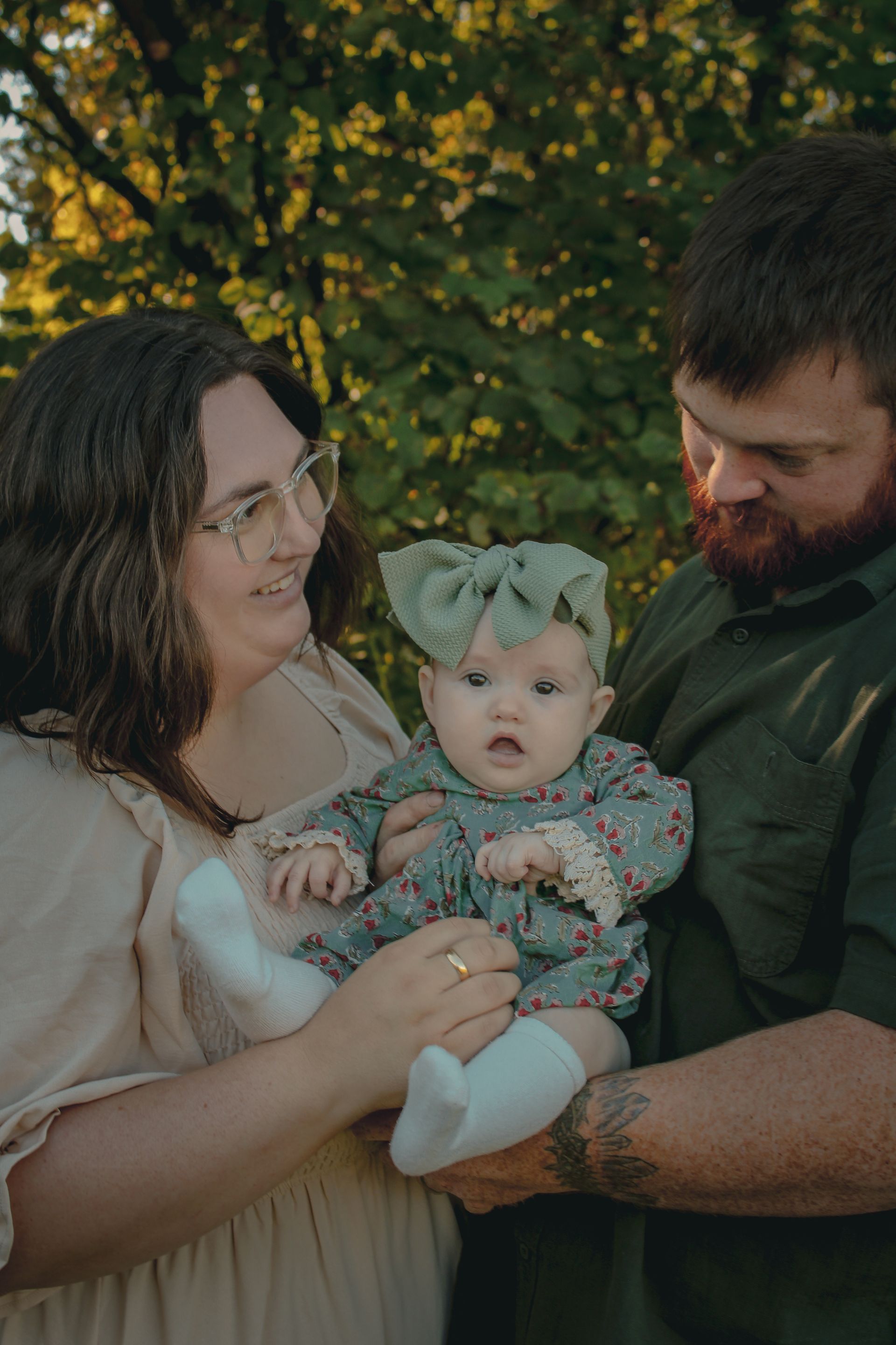 Adults holding a baby in a floral outfit outdoors, with trees in the background.