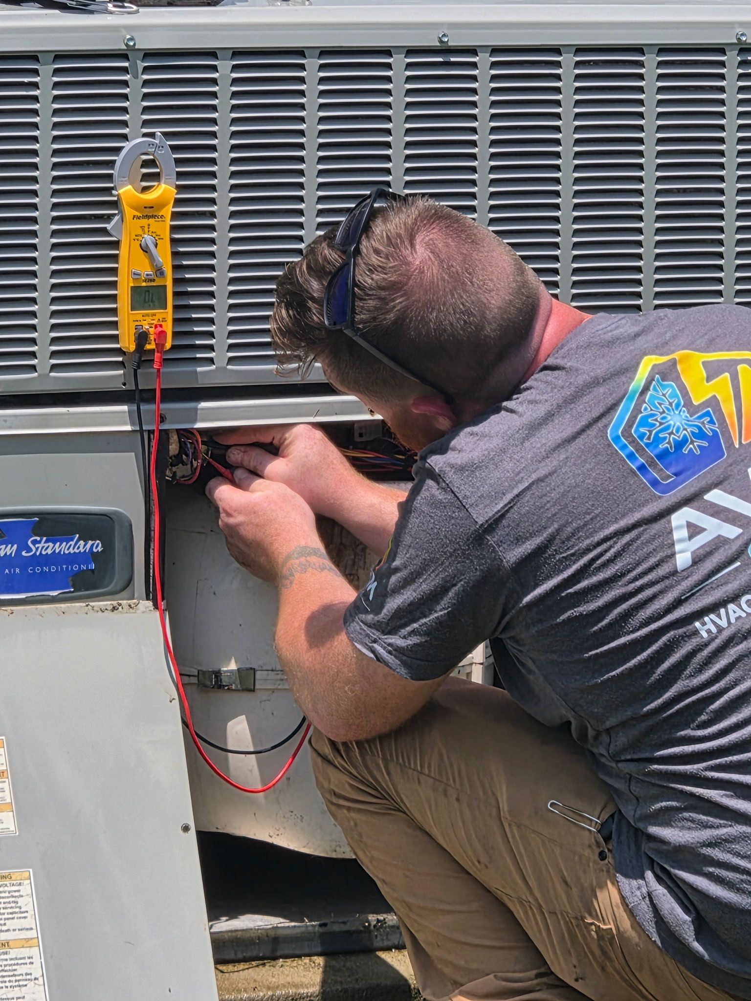 Technician kneeling beside HVAC unit, using a multimeter to inspect wiring and electrical components.