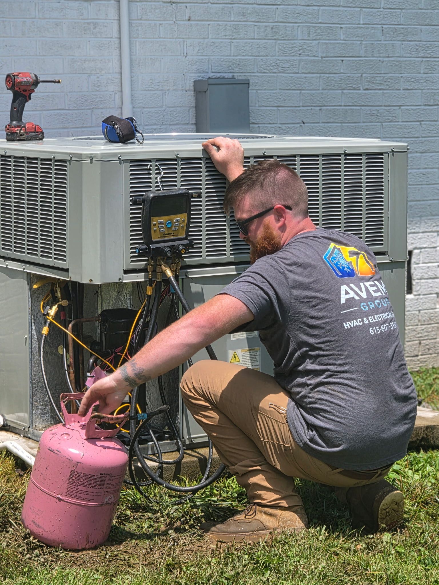 Technician kneels beside an HVAC unit, connecting a pink refrigerant tank outdoors.