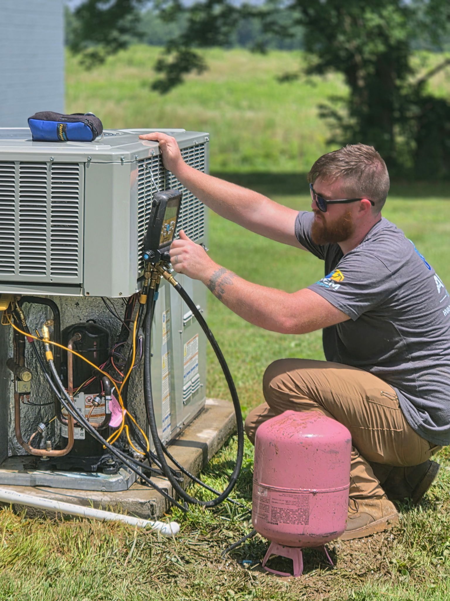 Technician kneels by an outdoor HVAC unit, adjusting wires beside a pink refrigerant tank.