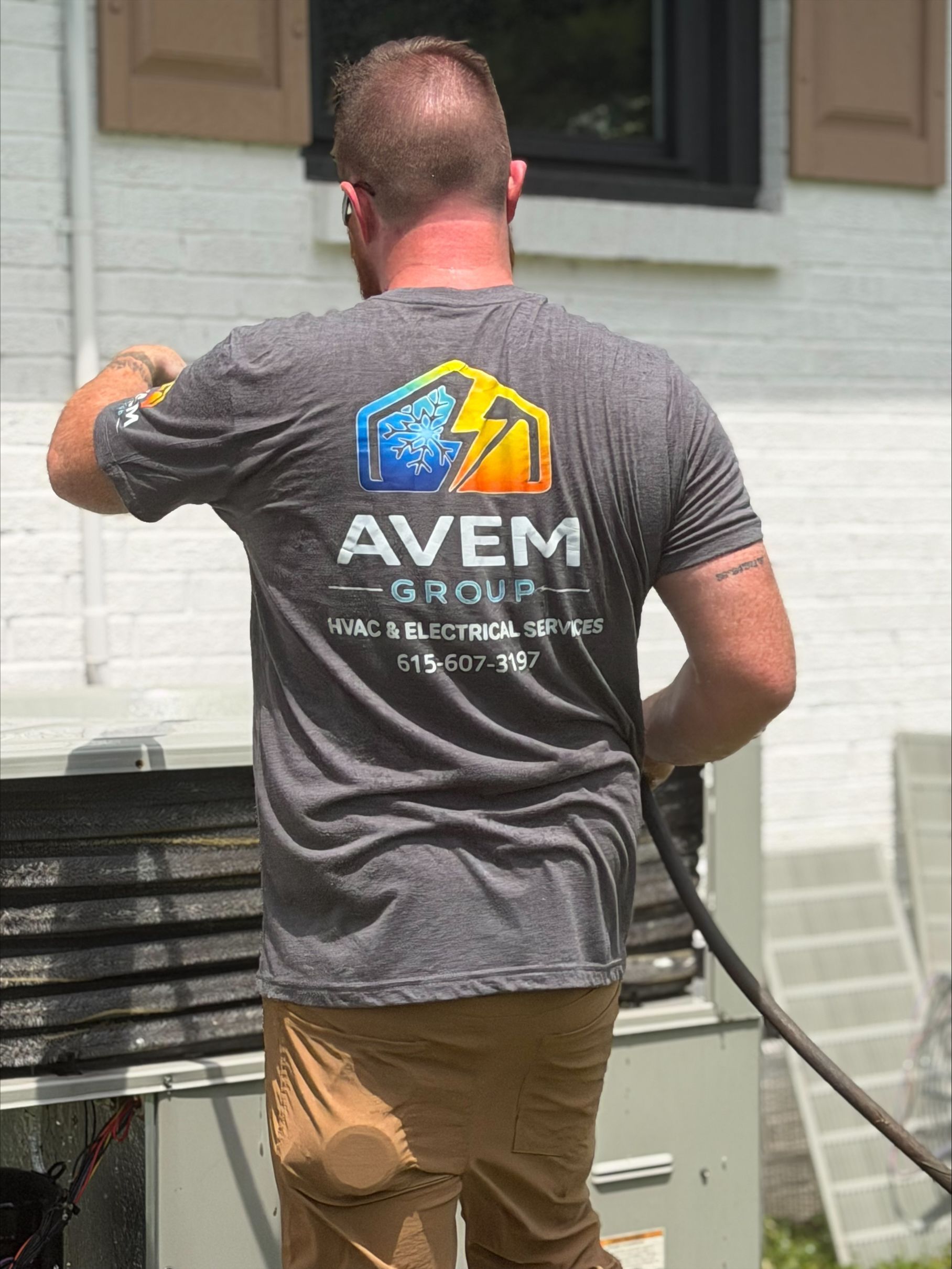 Man in gray AVEM shirt painting an exterior white wall near a window and AC unit
