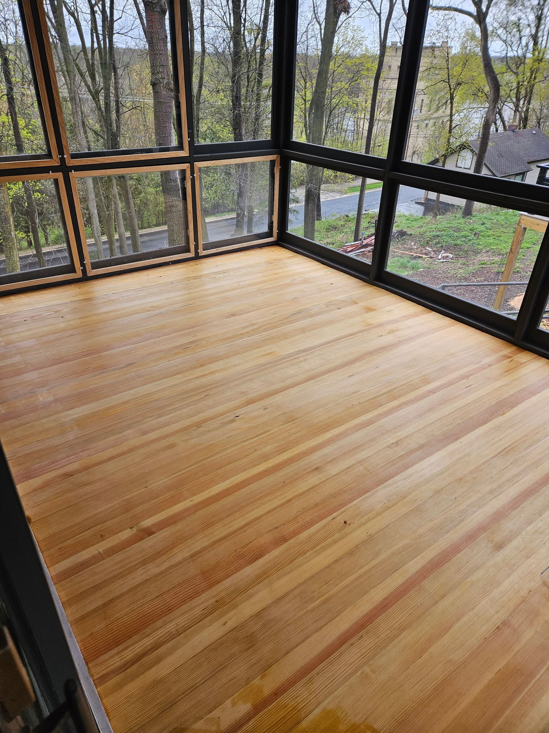 Wooden floor in a sunroom with large windows overlooking trees.
