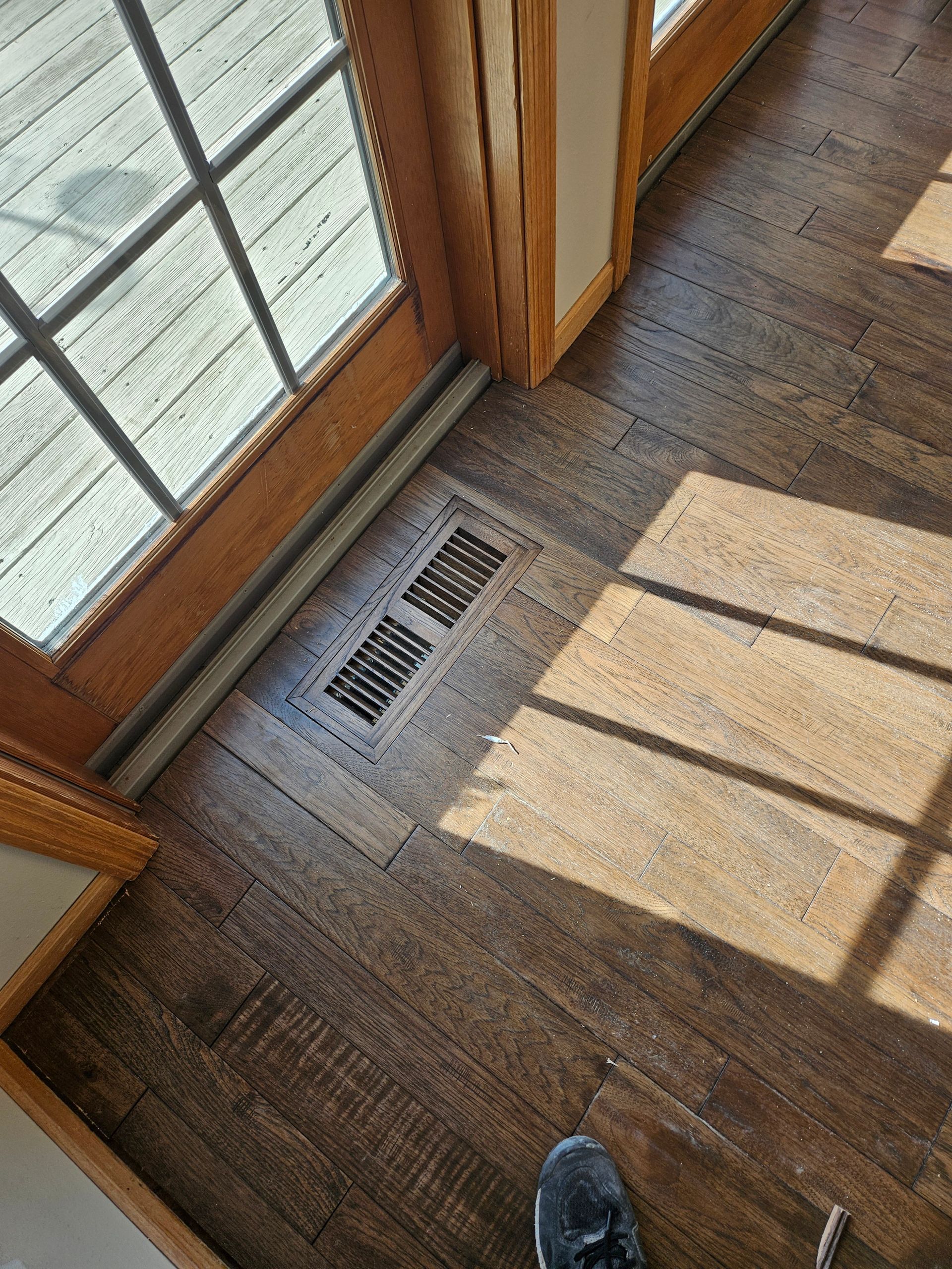 Hardwood floor with a vent near a glass door, sunlight casting shadows.