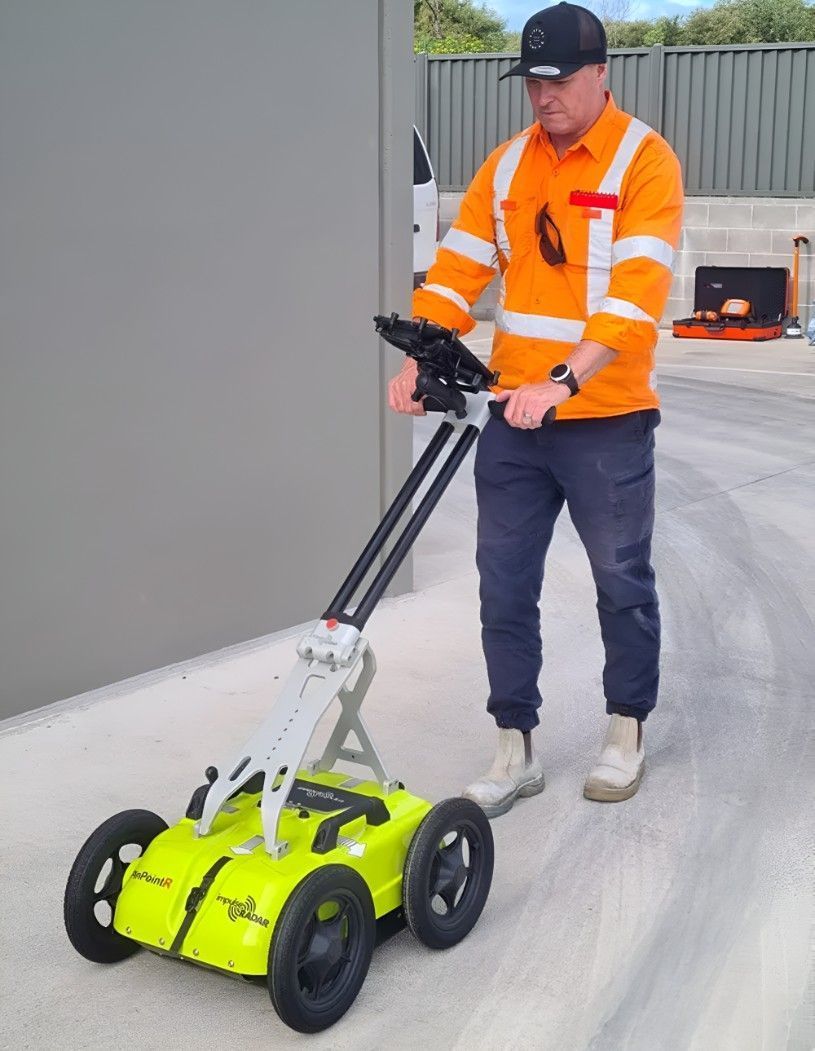 Person using a yellow and black ground-penetrating radar on wet asphalt, in an outdoor setting.