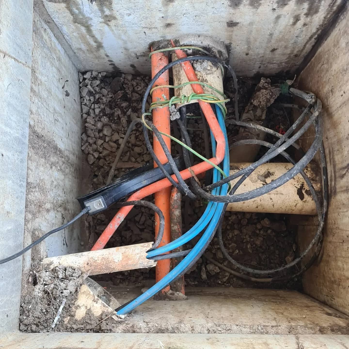 Inside a concrete box, orange and blue electrical conduits and black wires are visible, with rocks and debris.