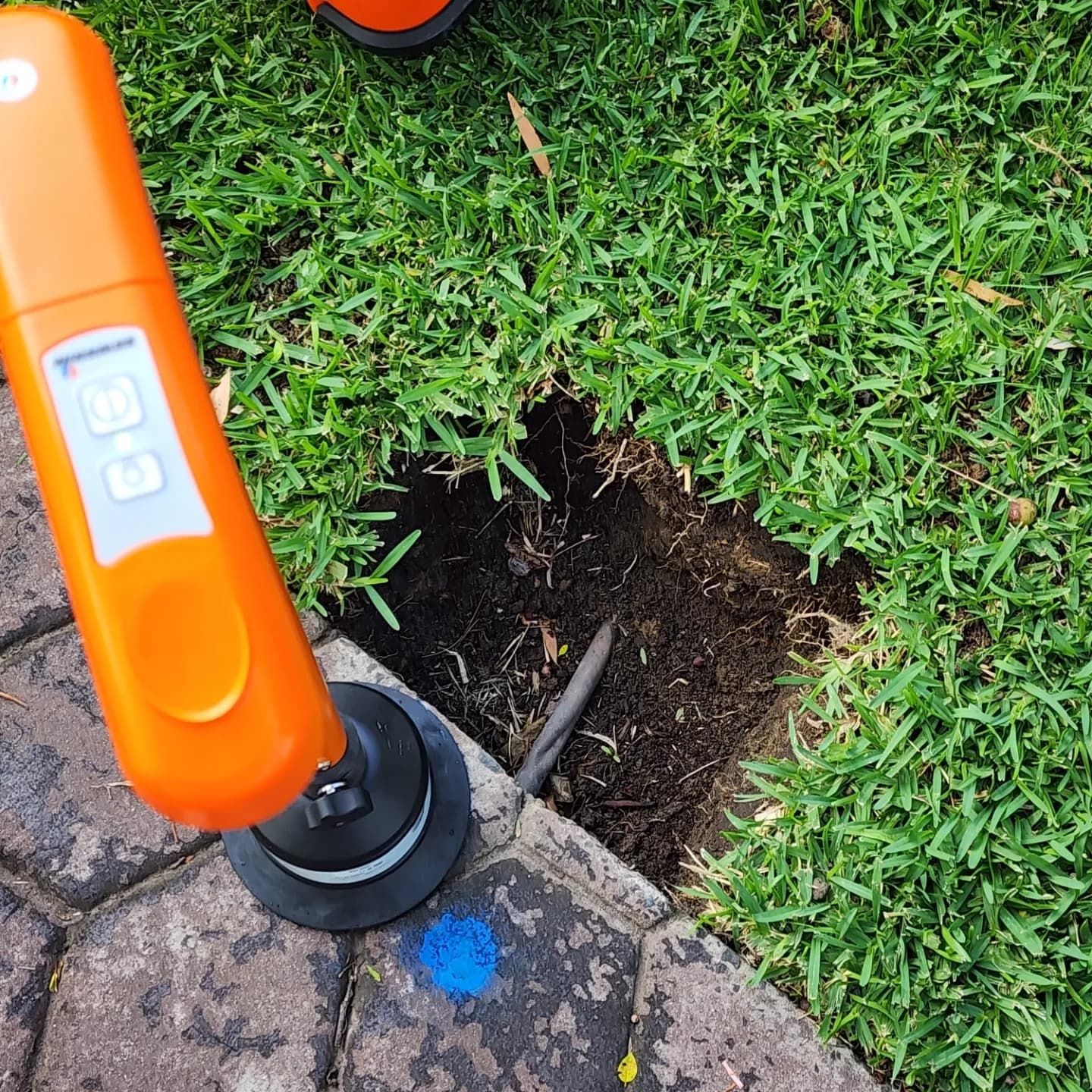 Orange utility locator next to a hole dug in grass near a brick edge.