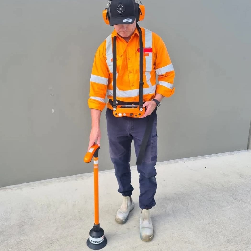 Person using an orange and black tool on the ground, wearing an orange safety vest, and black work boots.