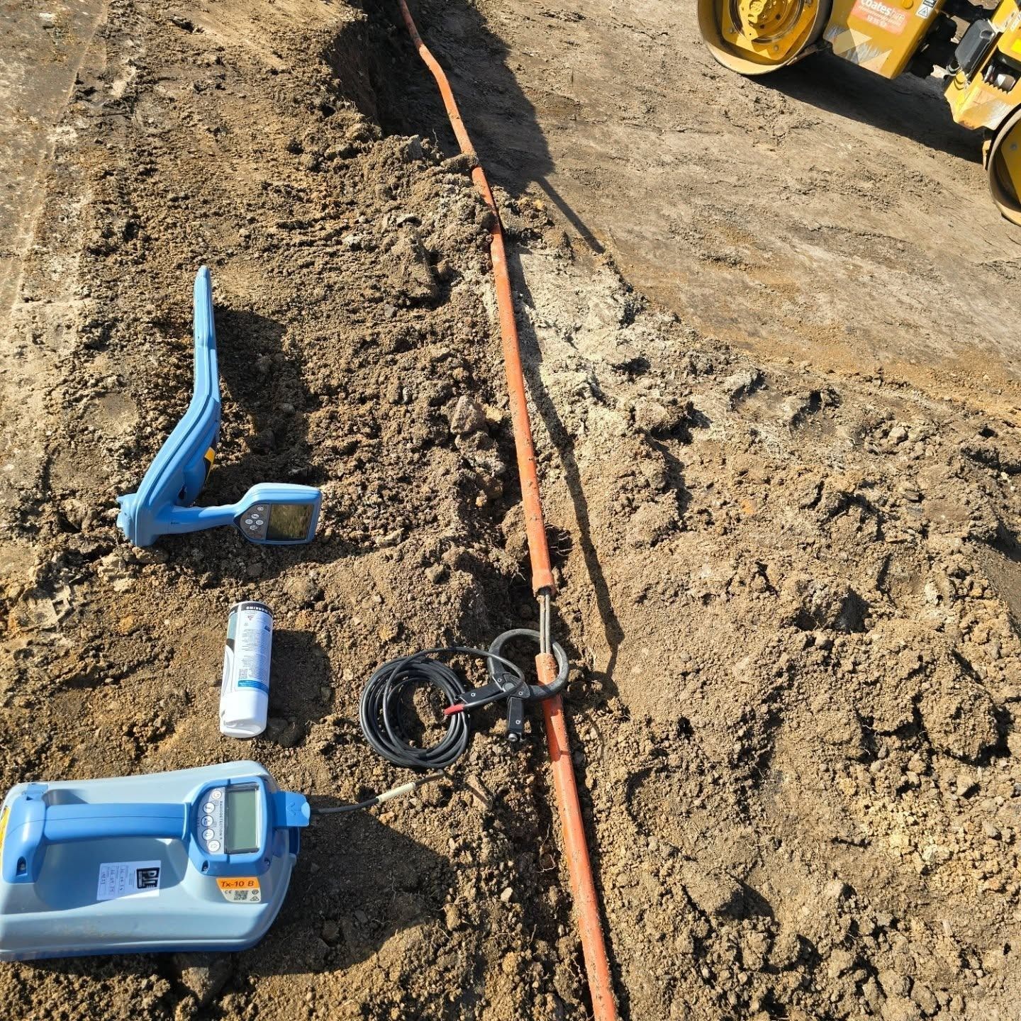 Construction site: A utility line in a trench with equipment like locators and a small excavator in the background.