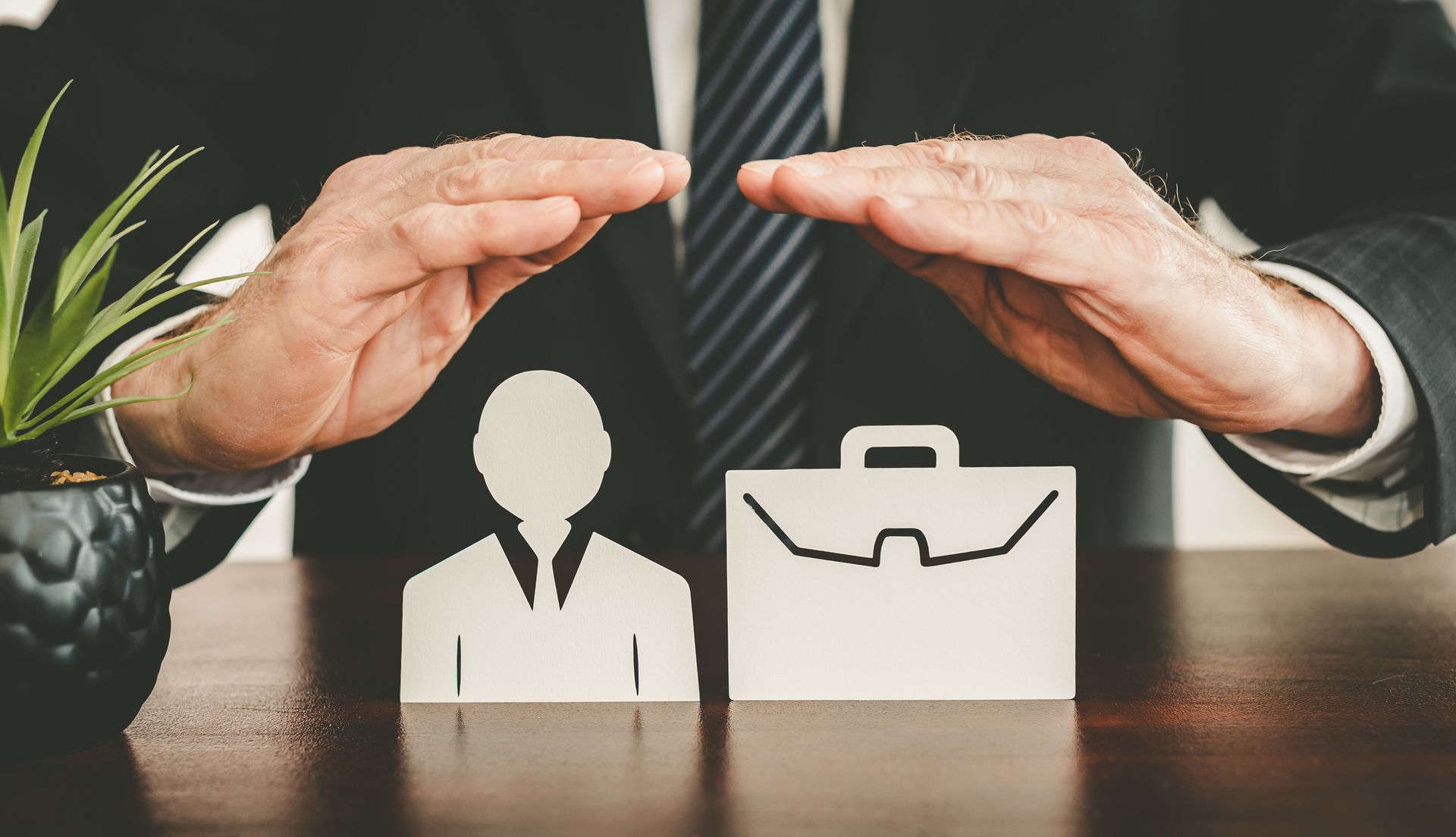Hands sheltering icons of a person and briefcase on a desk, symbolizing protection of jobs.