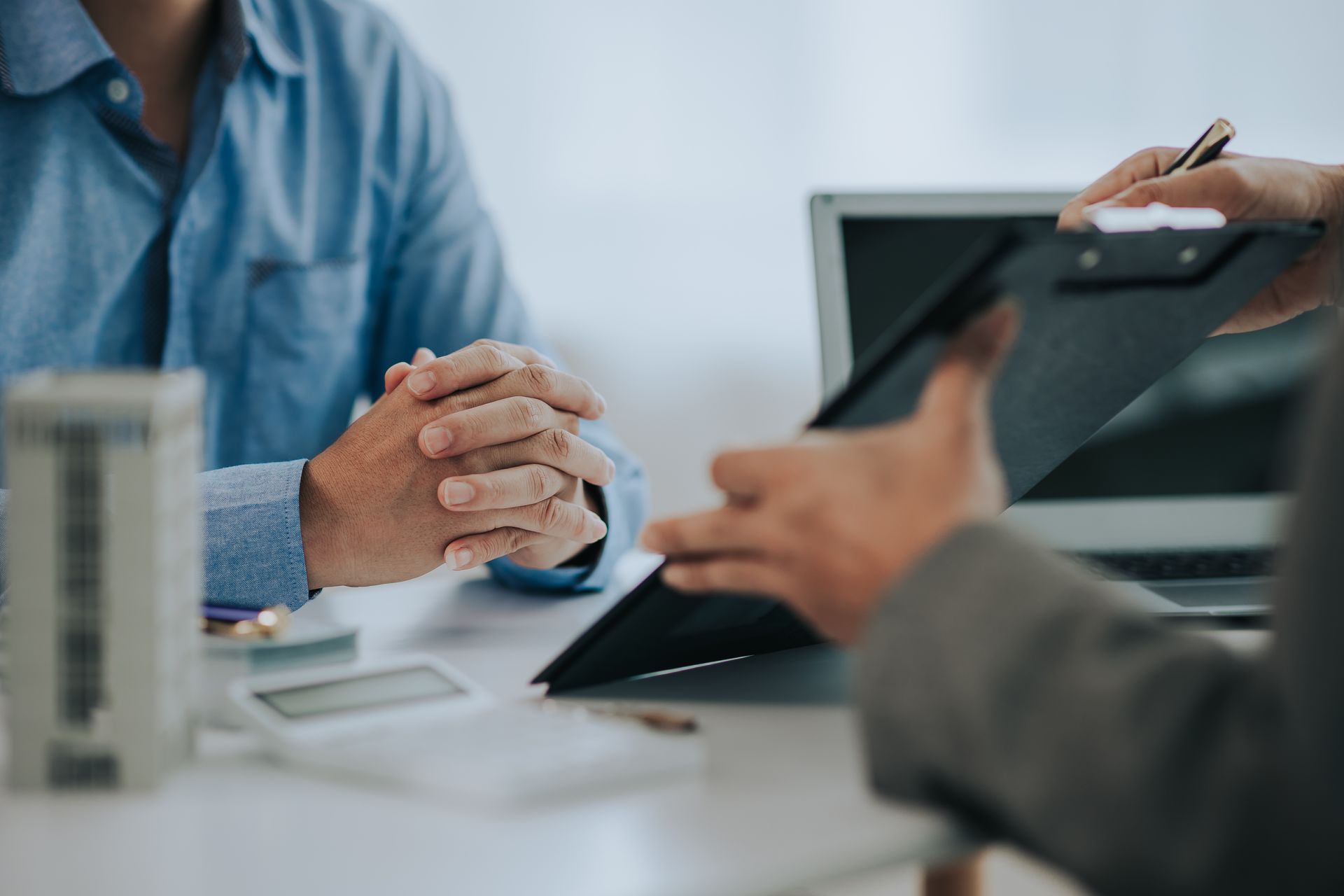 Two people in a business meeting, one holding a tablet and one clasping hands at a desk.