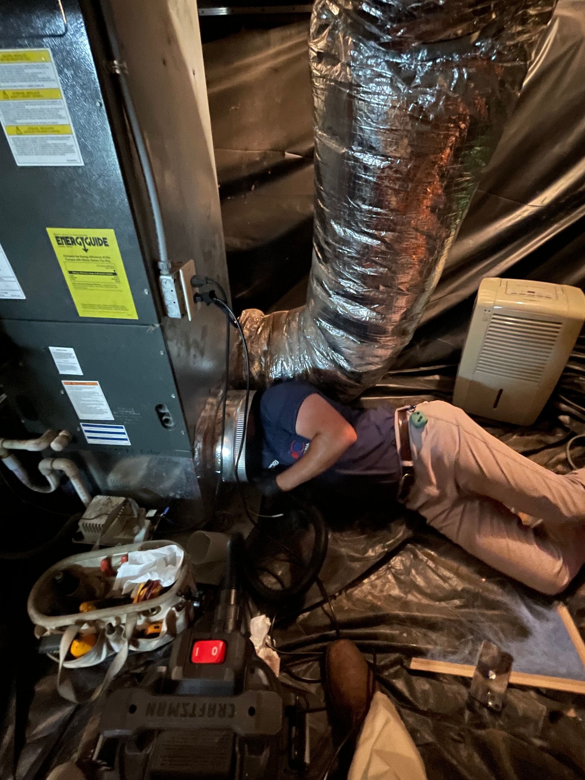 Person working on HVAC ductwork in a crawl space. They are wearing light pants and a blue shirt, surrounded by tools and equipment.
