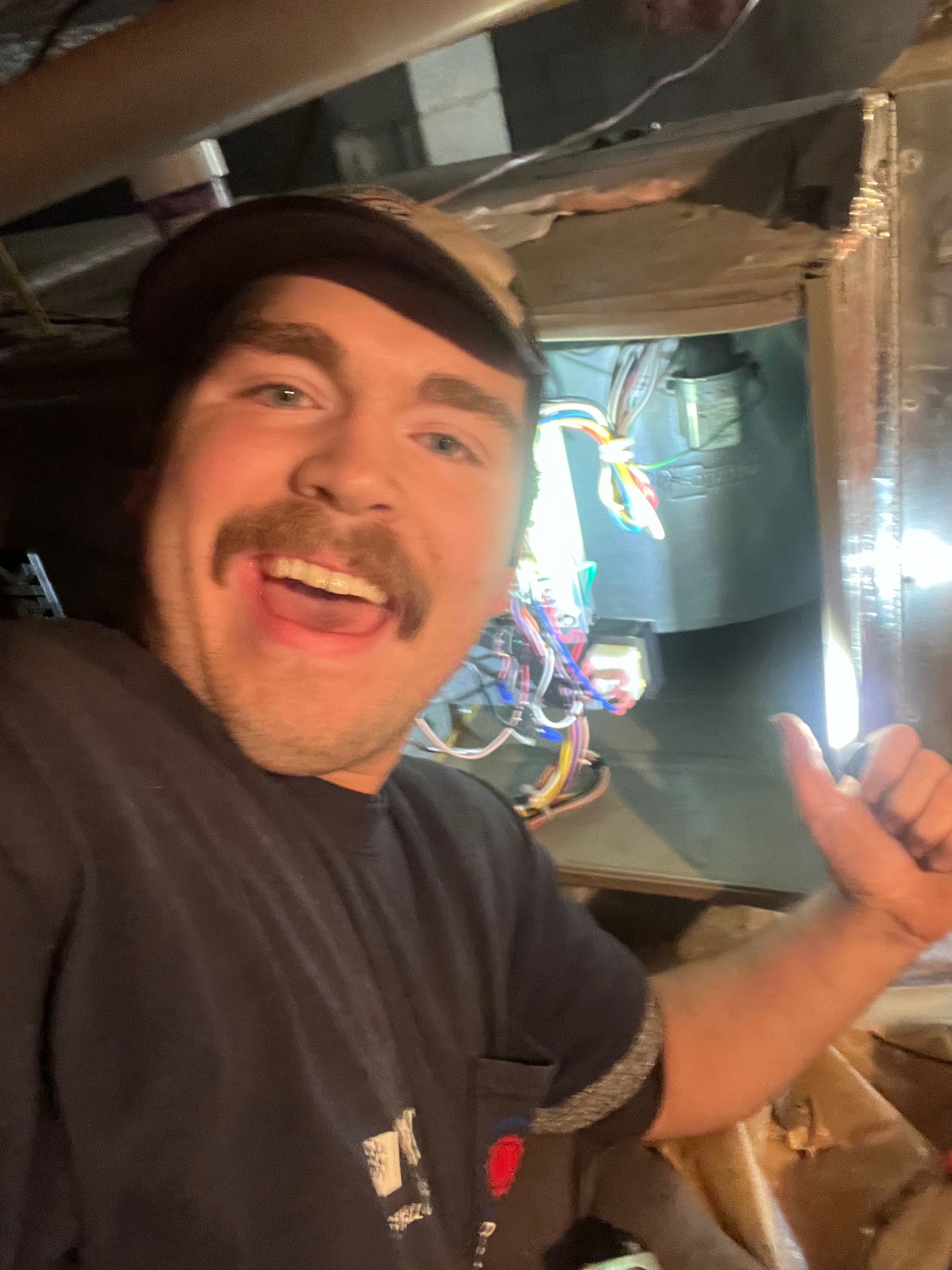 Man with mustache giving a thumbs-up, working on wires in a tight space.