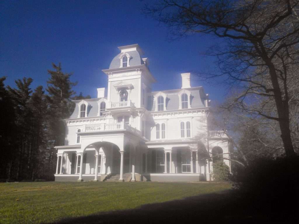 White Victorian mansion with gray roof and trim on a sunny day, surrounded by trees and lawn.