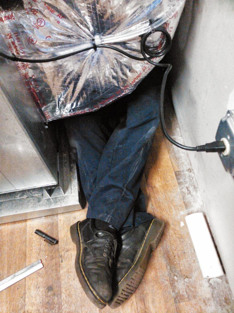 Person lying down with legs crossed, inside of a metal duct. Wearing black boots and jeans, power cord nearby.