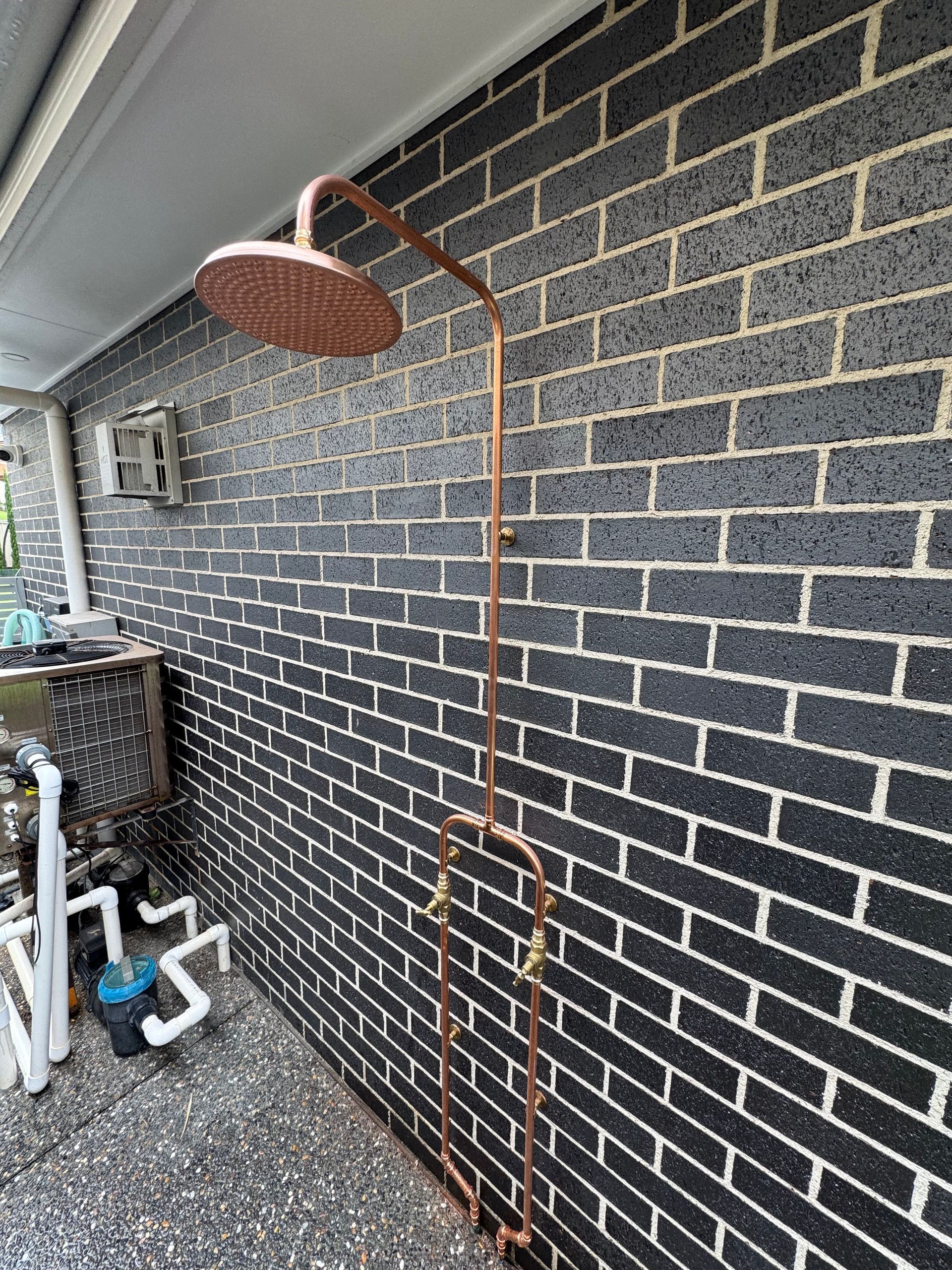 Copper outdoor shower head and pipes on a black brick wall — Fair Plumbing In Shellharbour, NSW