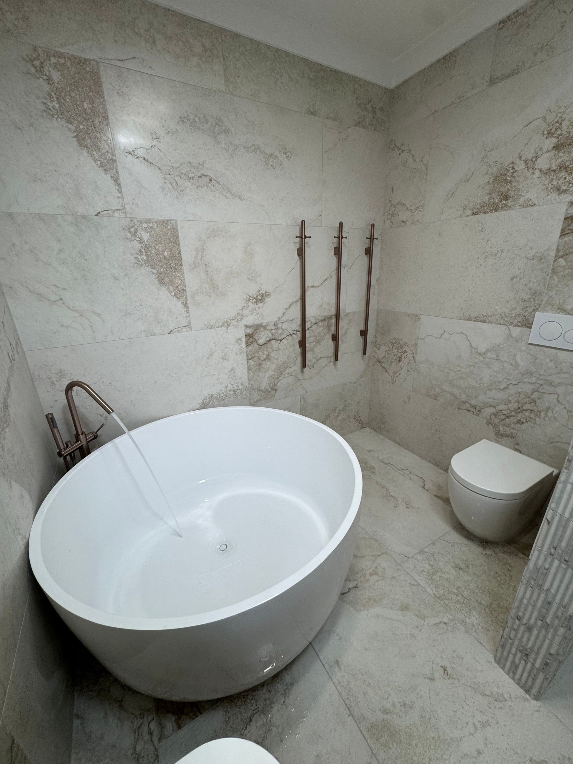 Modern bathroom with white oval tub, toilet, and beige marble tile. Copper fixtures  — Fair Plumbing In Shellharbour, NSW