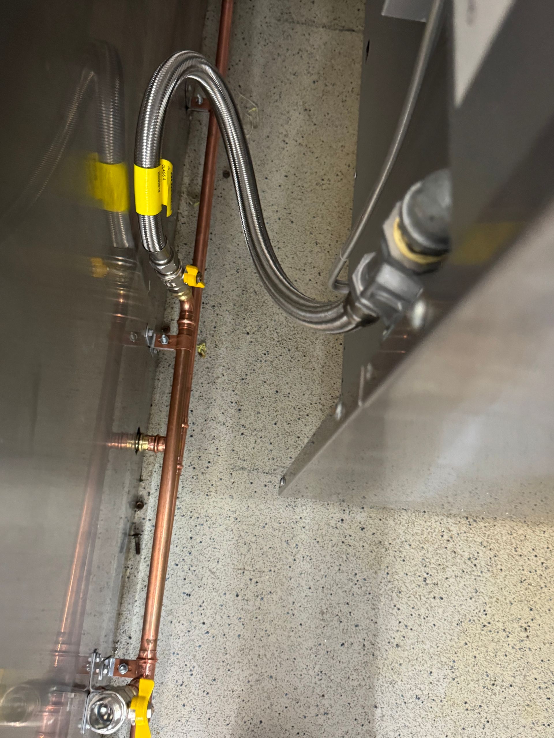 Copper pipes and a flexible hose connected to a silver appliance, secured with yellow clamps, set against a speckled wall — Fair Plumbing In Shellharbour, NSW