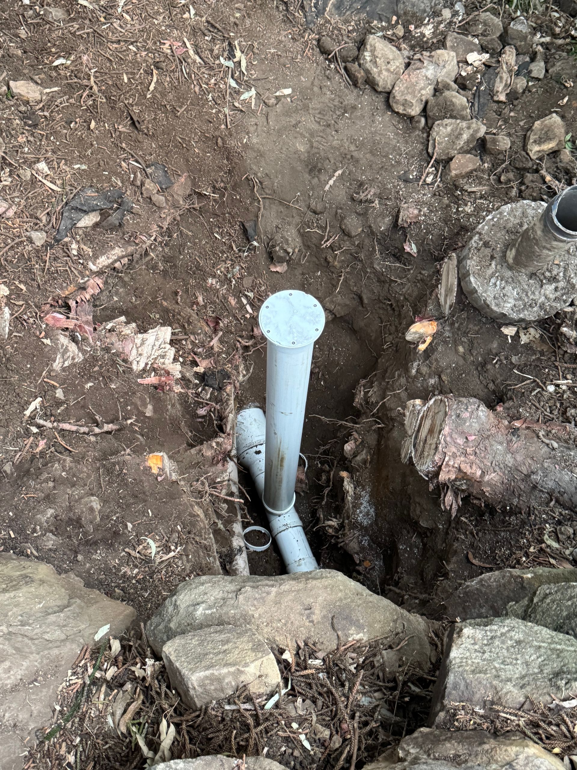 Pipes in a dirt trench, rocks on the sides. One pipe has a gray cap  — Fair Plumbing In Shellharbour, NSW